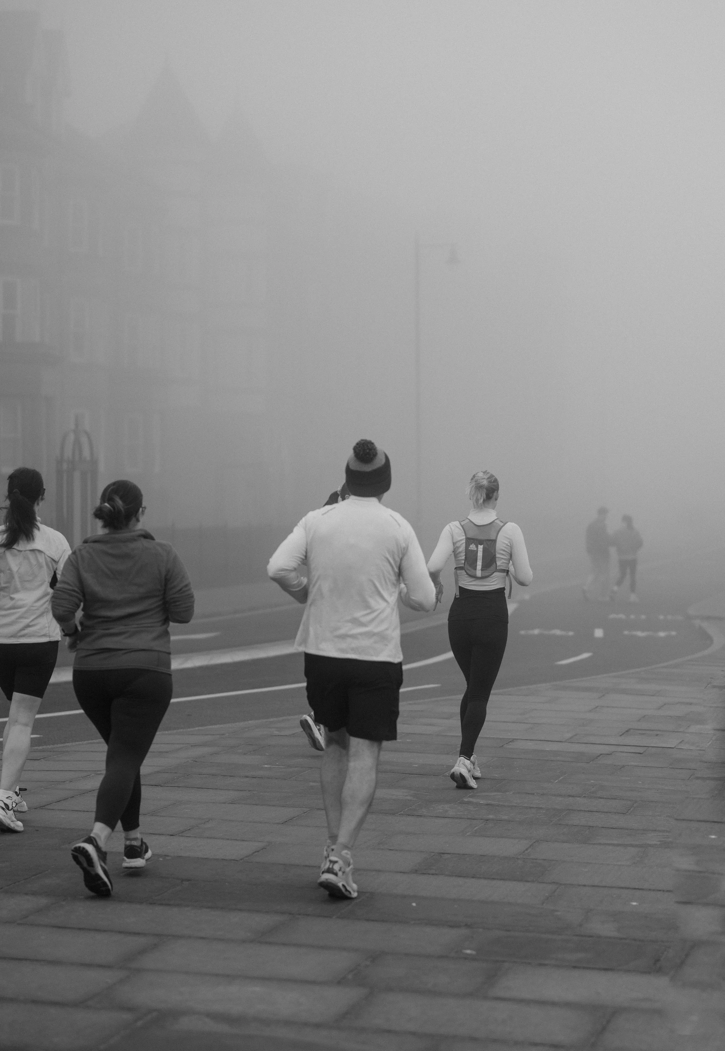 People jogging on sidewalk in foggy weather, some wearing jackets and athletic gear.