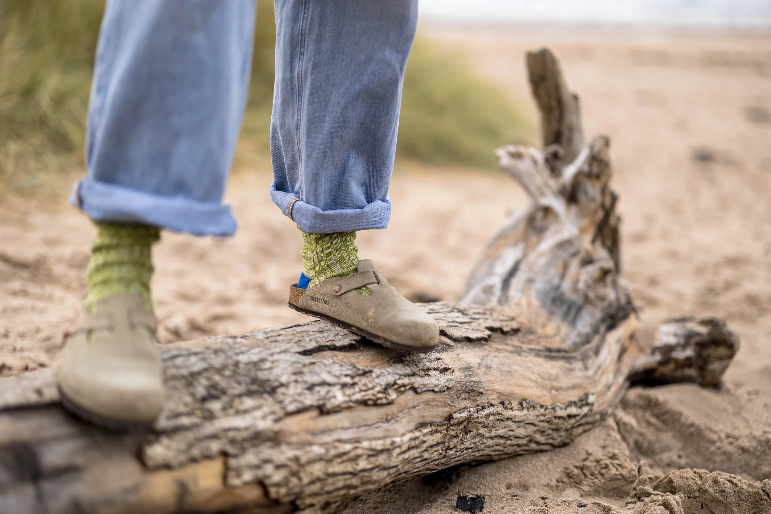 Person standing on a piece of driftwood on a sandy beach, wearing rolled-up jeans, green socks, and beige shoes.
