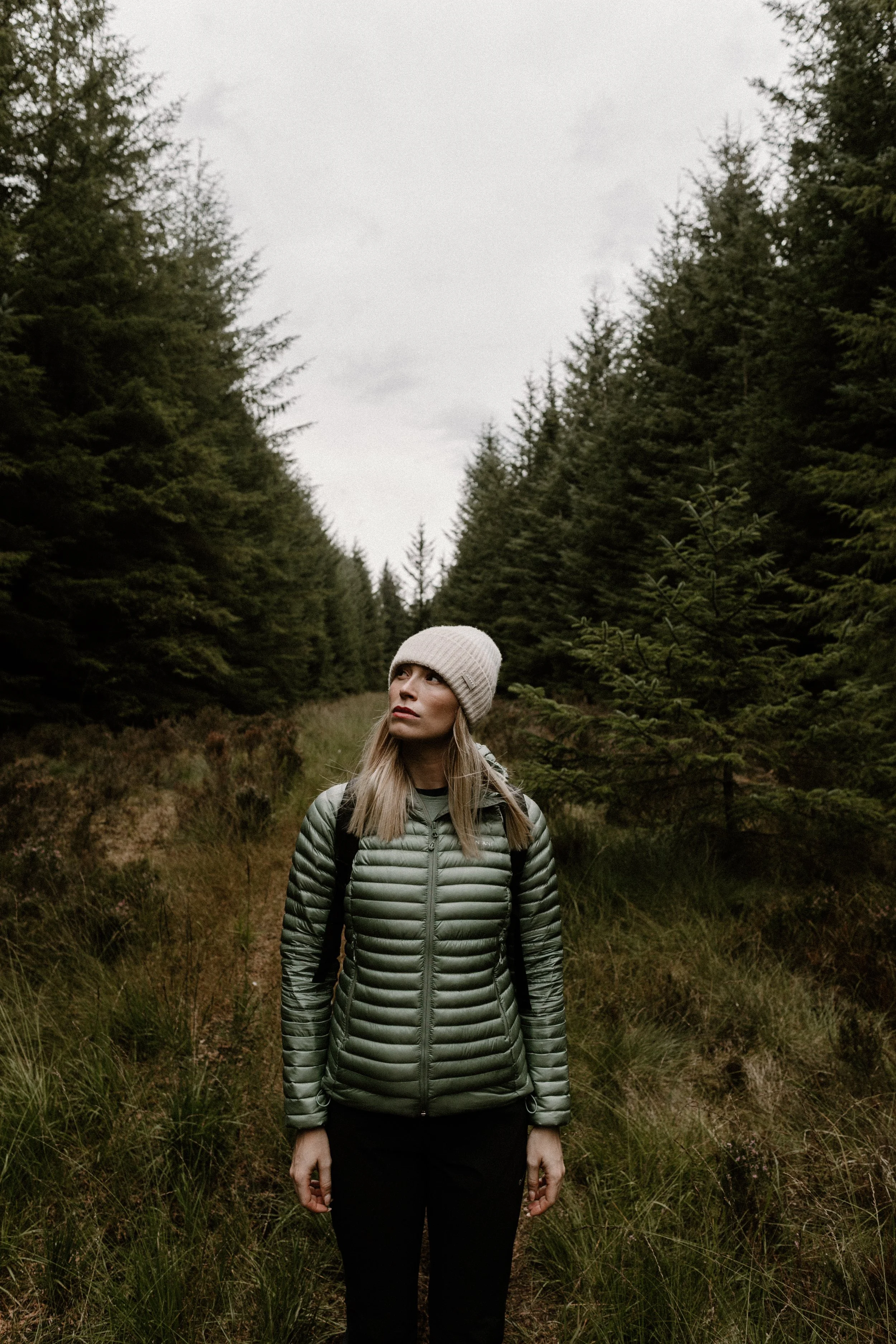 A woman in a green puffer jacket and beige beanie standing in a forested area with tall evergreen trees.
