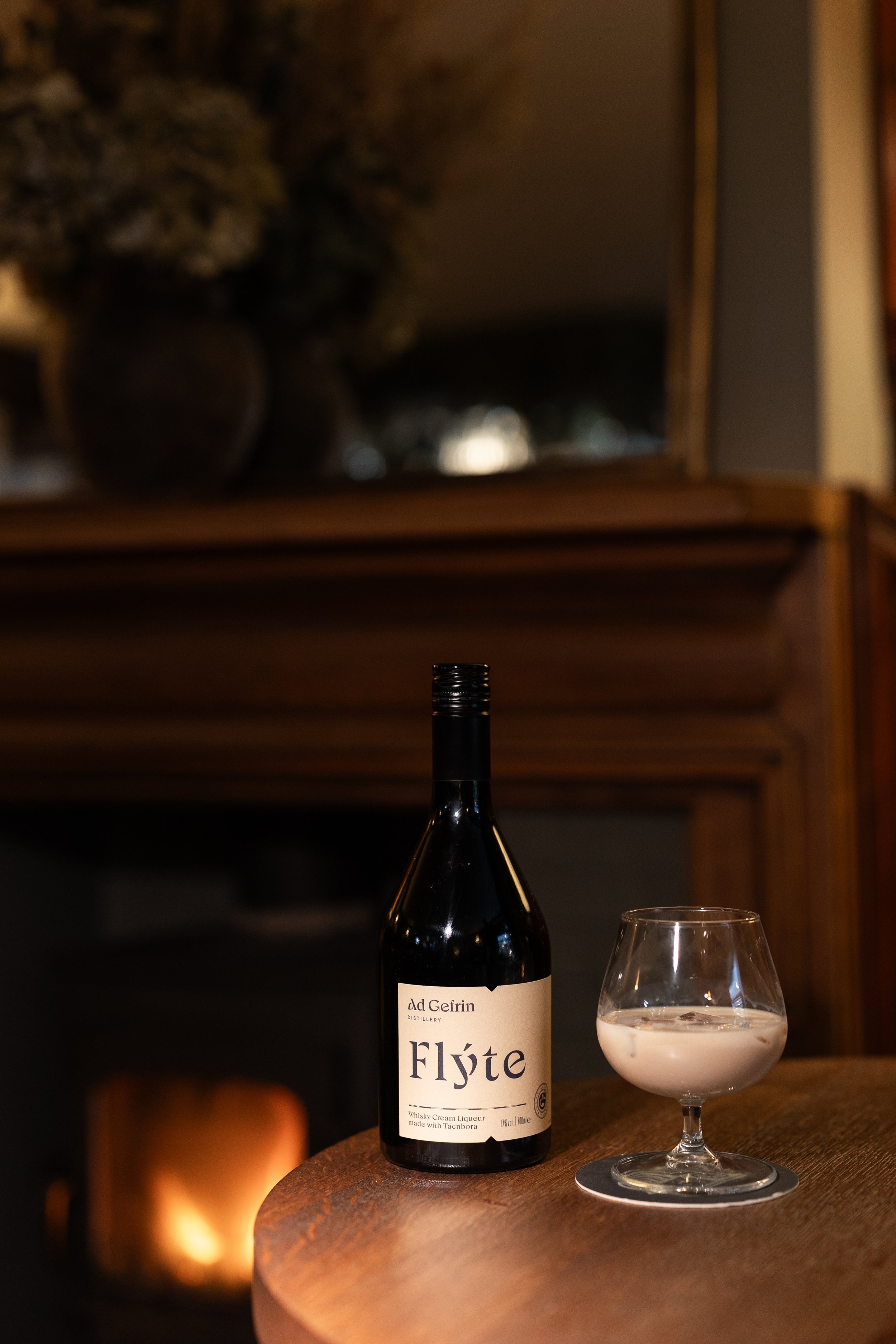 Bottle of Gefrin Flyte whisky cream liqueur and a glass of the drink on a wooden surface in a cozy room.