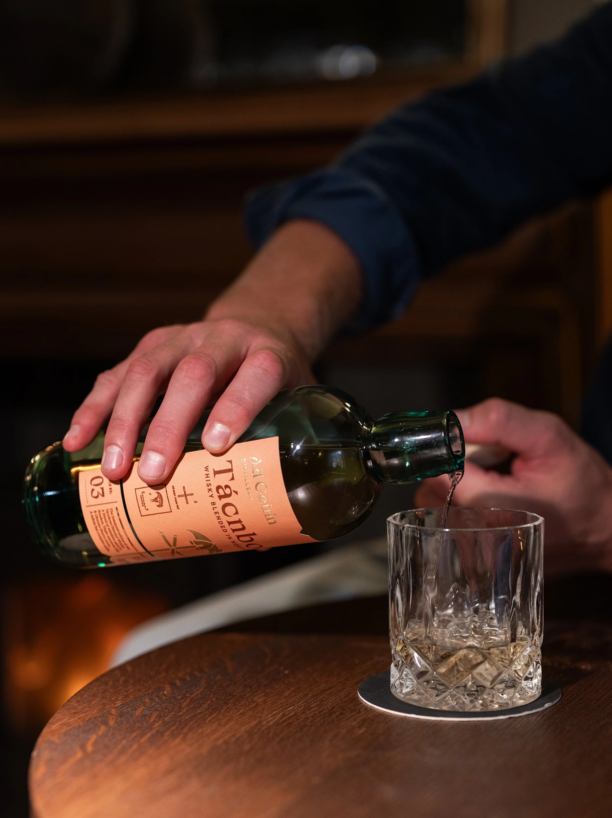 Person pouring whiskey into a glass on a wooden table.