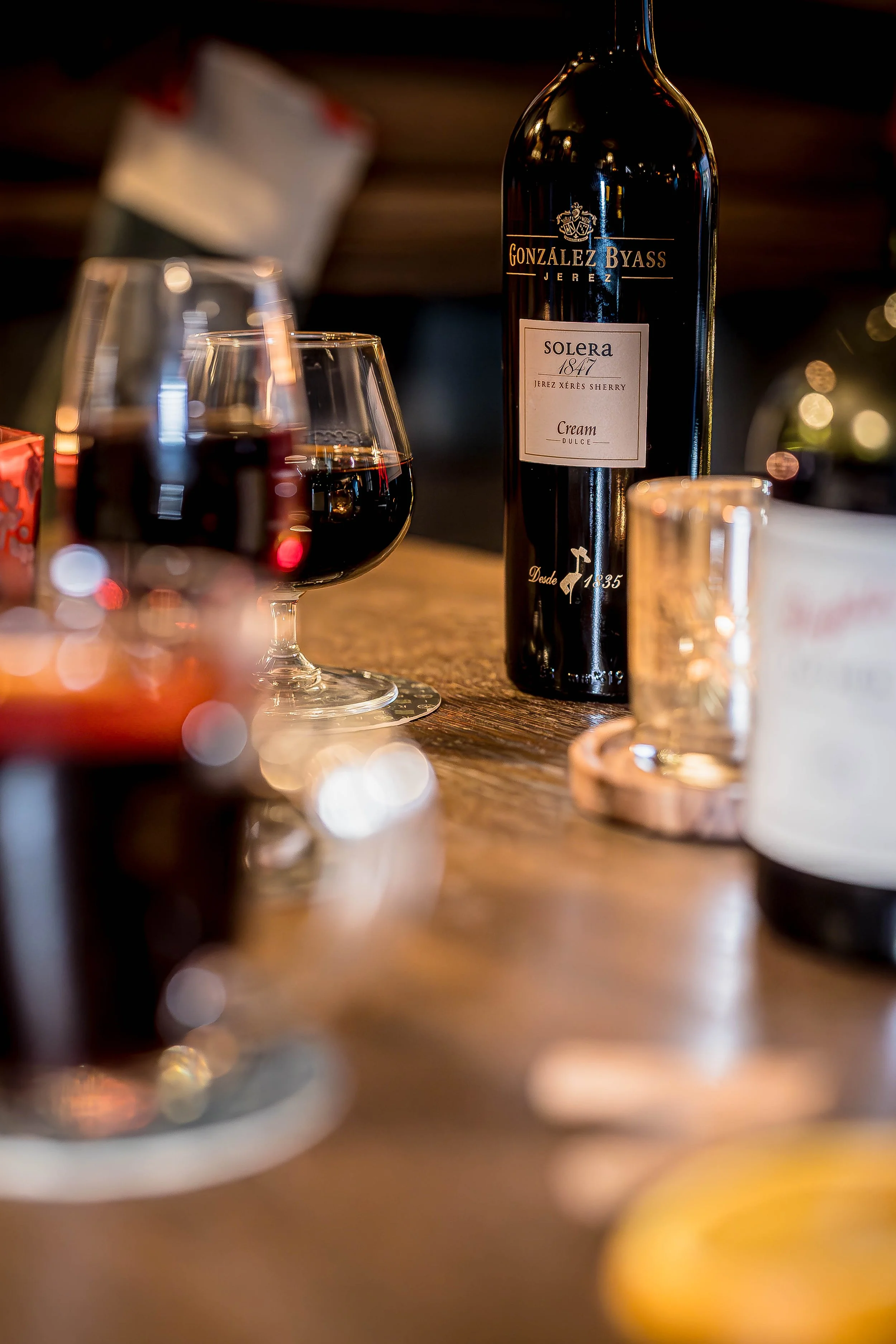A wine bottle labeled 'González Byass Solera 1847' on a wooden table with two glasses of red wine, some blurry objects, and a cozy, dimly-lit background.