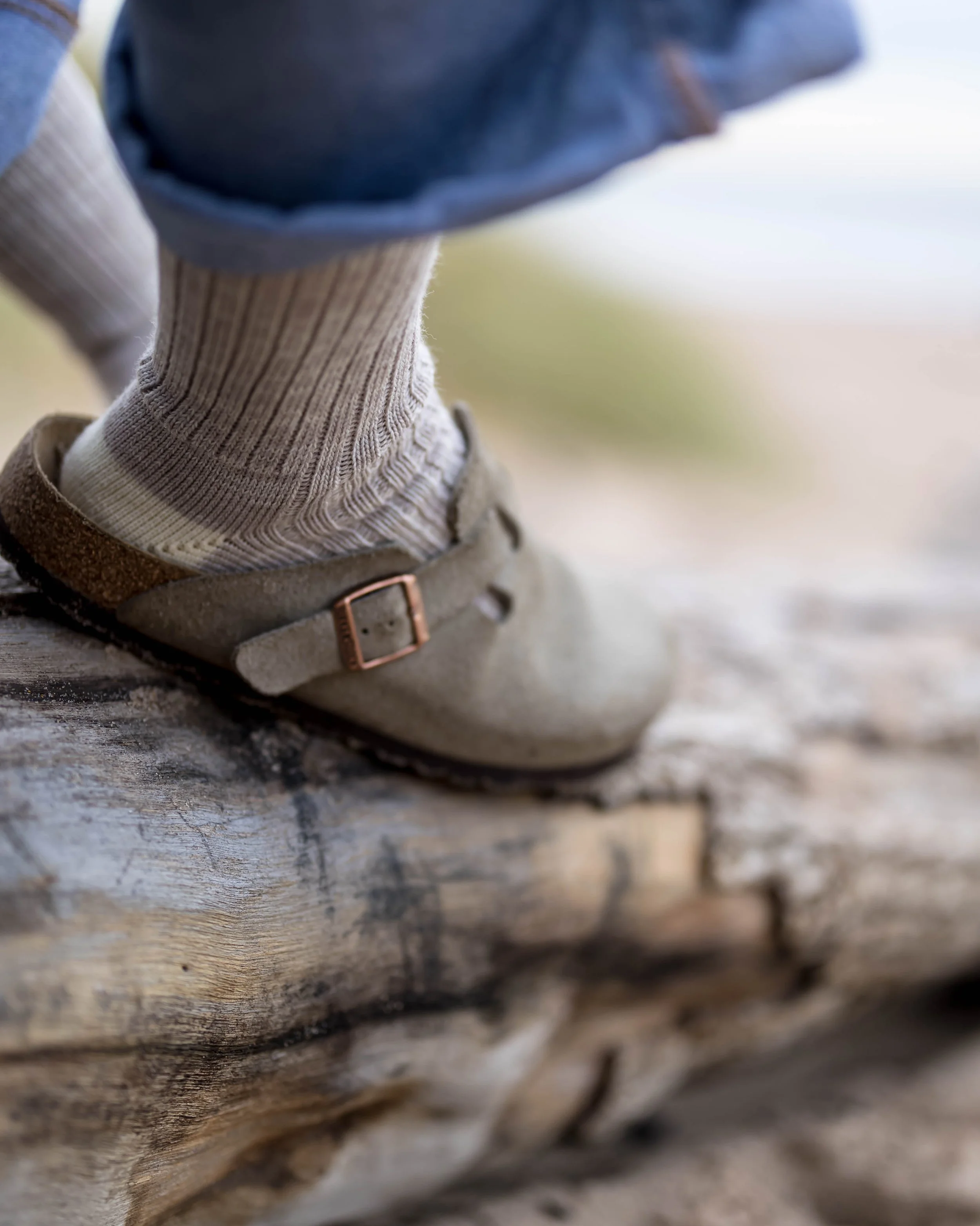 Close-up of a child's foot wearing a beige suede shoe with a strap and metal buckle, beige socks, blue jeans, and standing on a weathered wooden log.