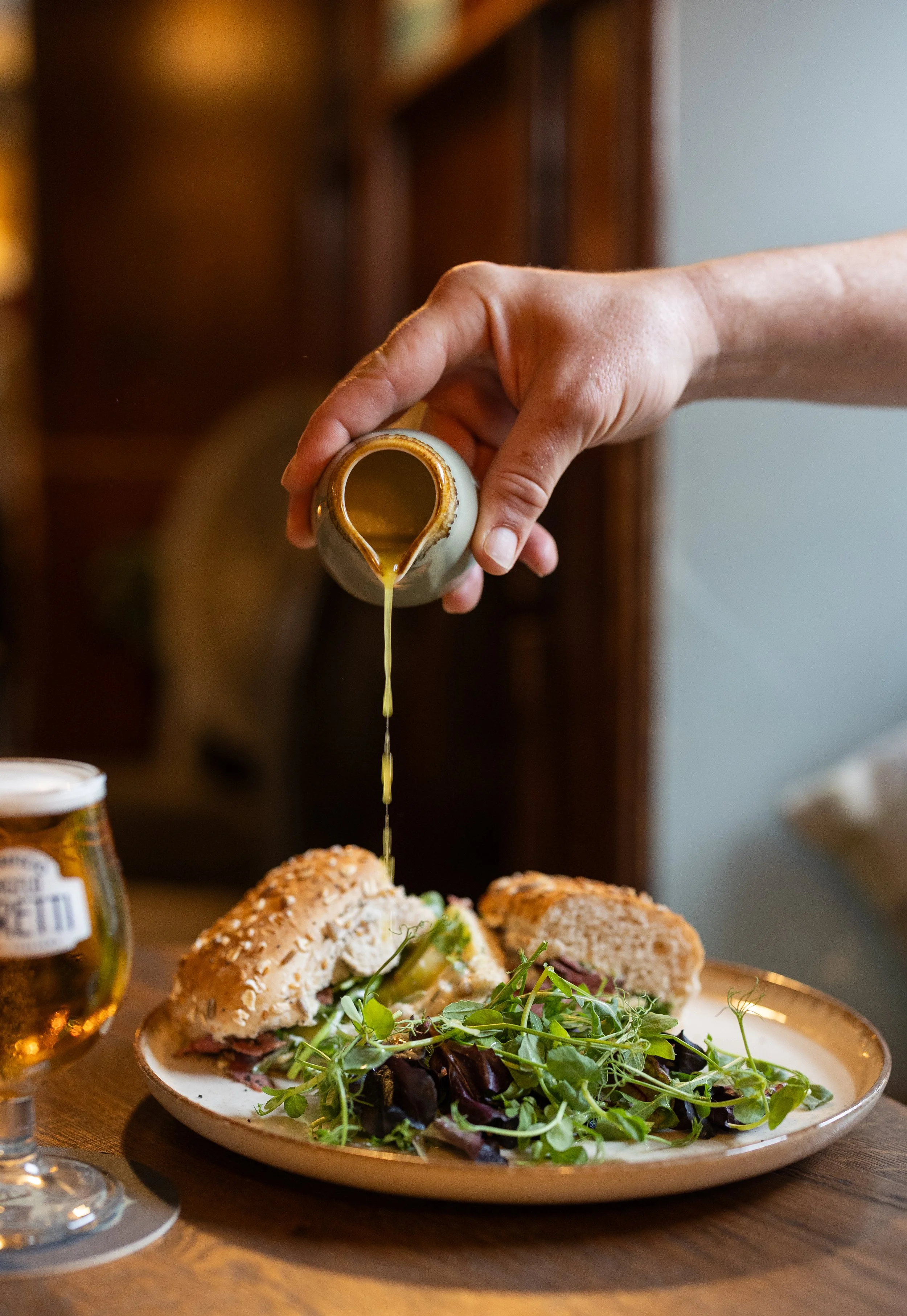 A hand pouring dressing on a sandwich with greens on a plate, next to a glass of beer.