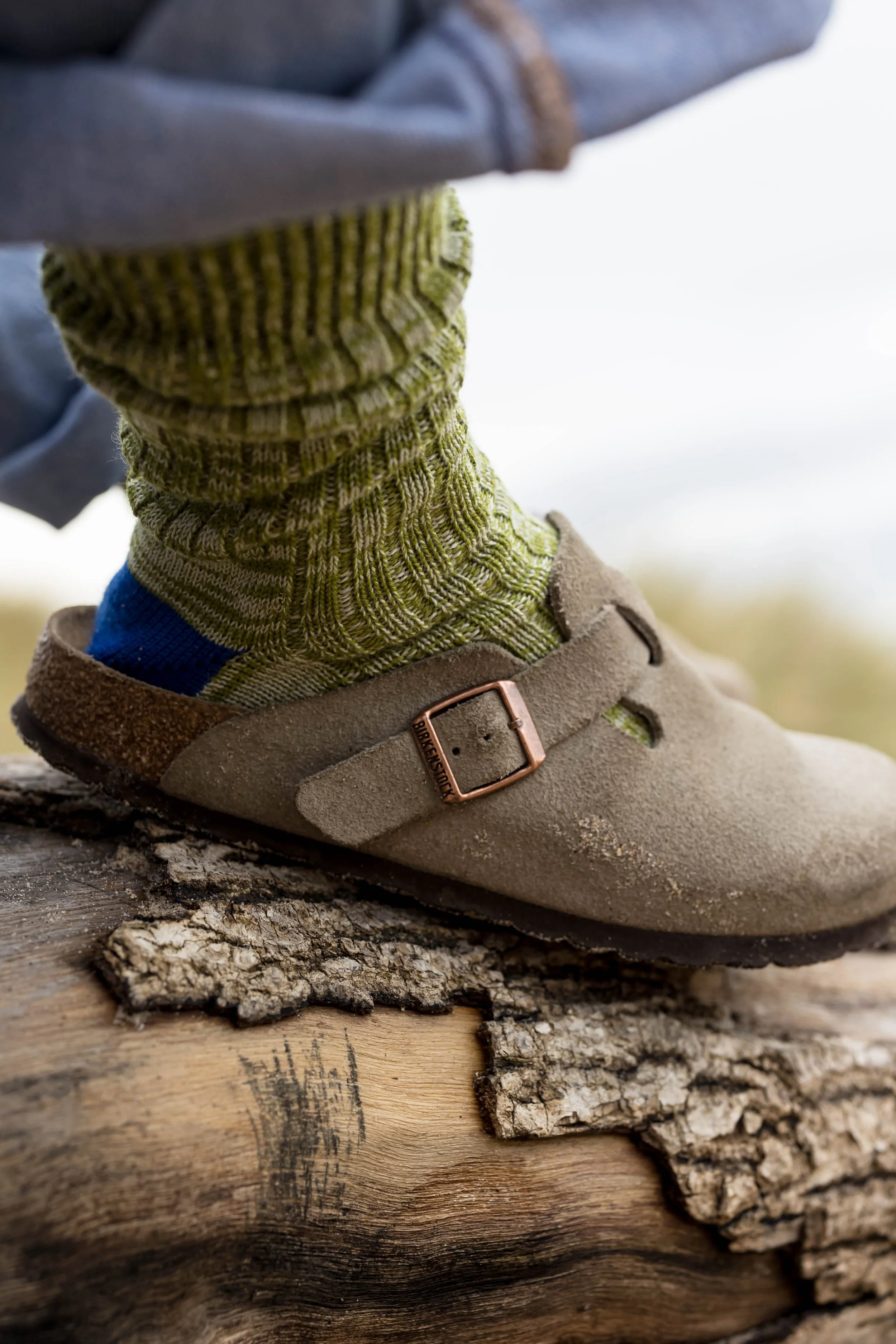 Close-up of a person's foot wearing a brown suede clog with a strap and buckle, green knit sock, blue athletic sock, and gray pants, standing on a rough log outdoors.
