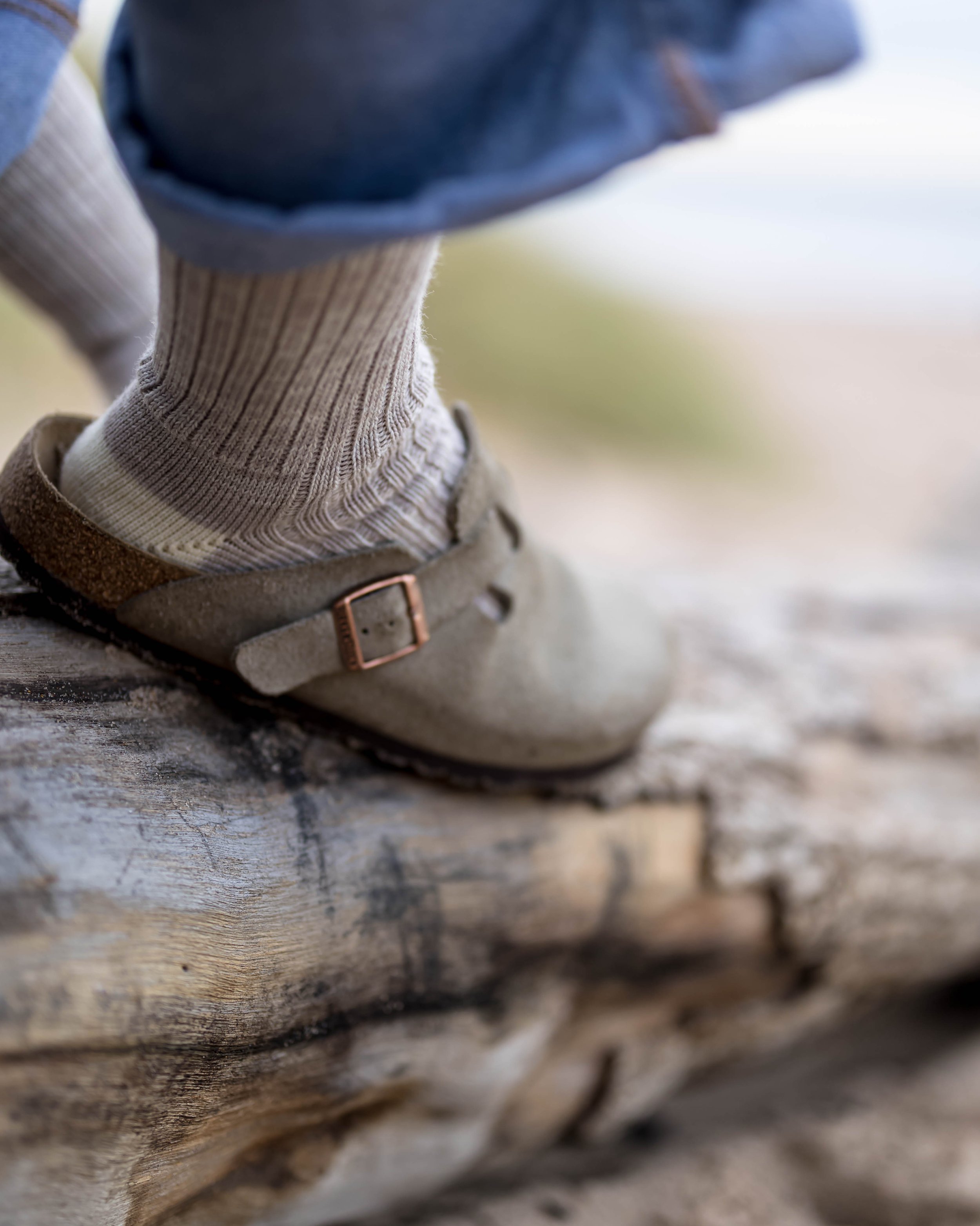 Close-up of a child's foot in a beige boot with a bronze buckle, wearing a gray sock, stepping on weathered wood outdoors.