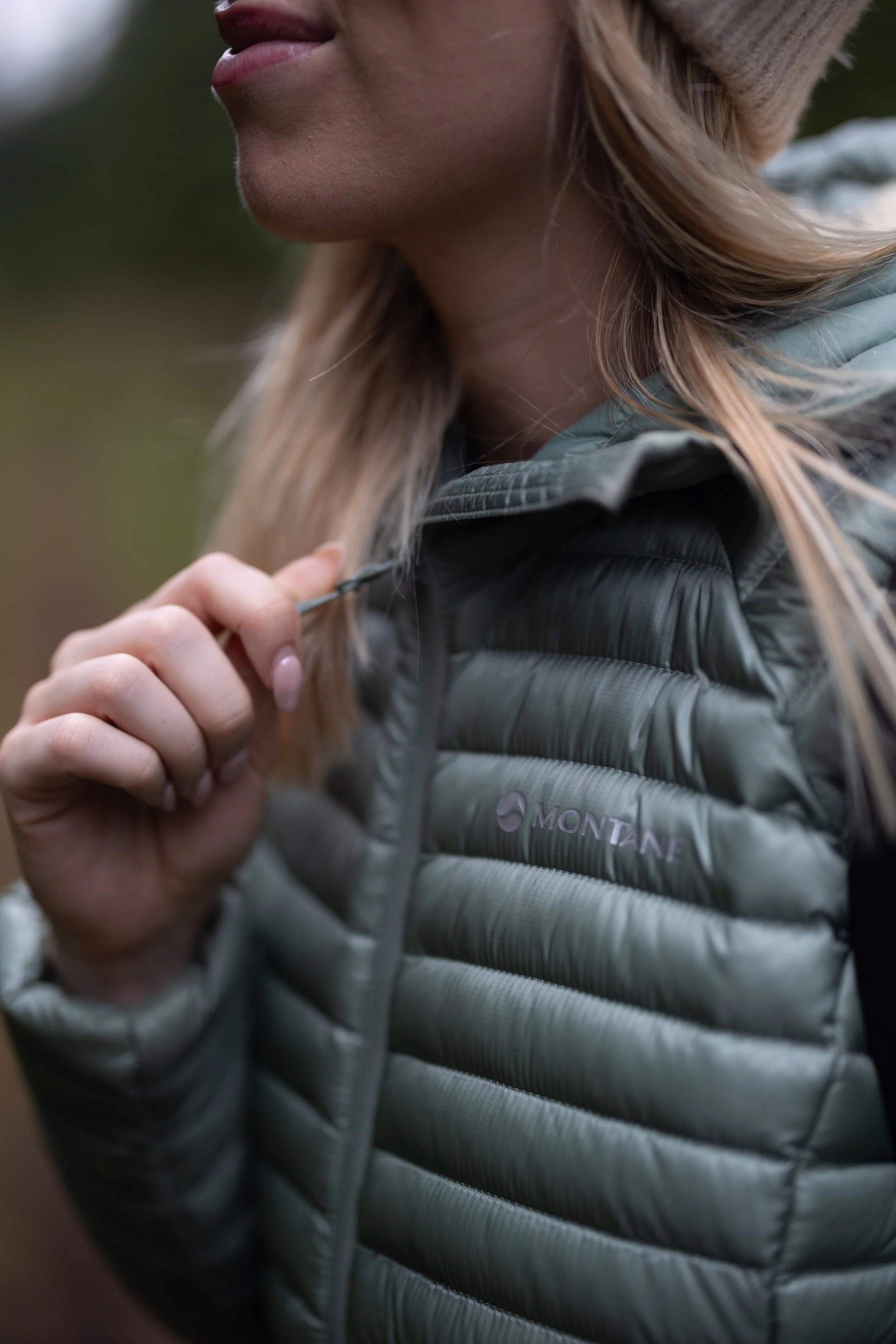 Close-up of a woman in a green puffer jacket with the brand 'Montane' logo, holding the jacket's zipper with her hand, outdoors with blurred background.