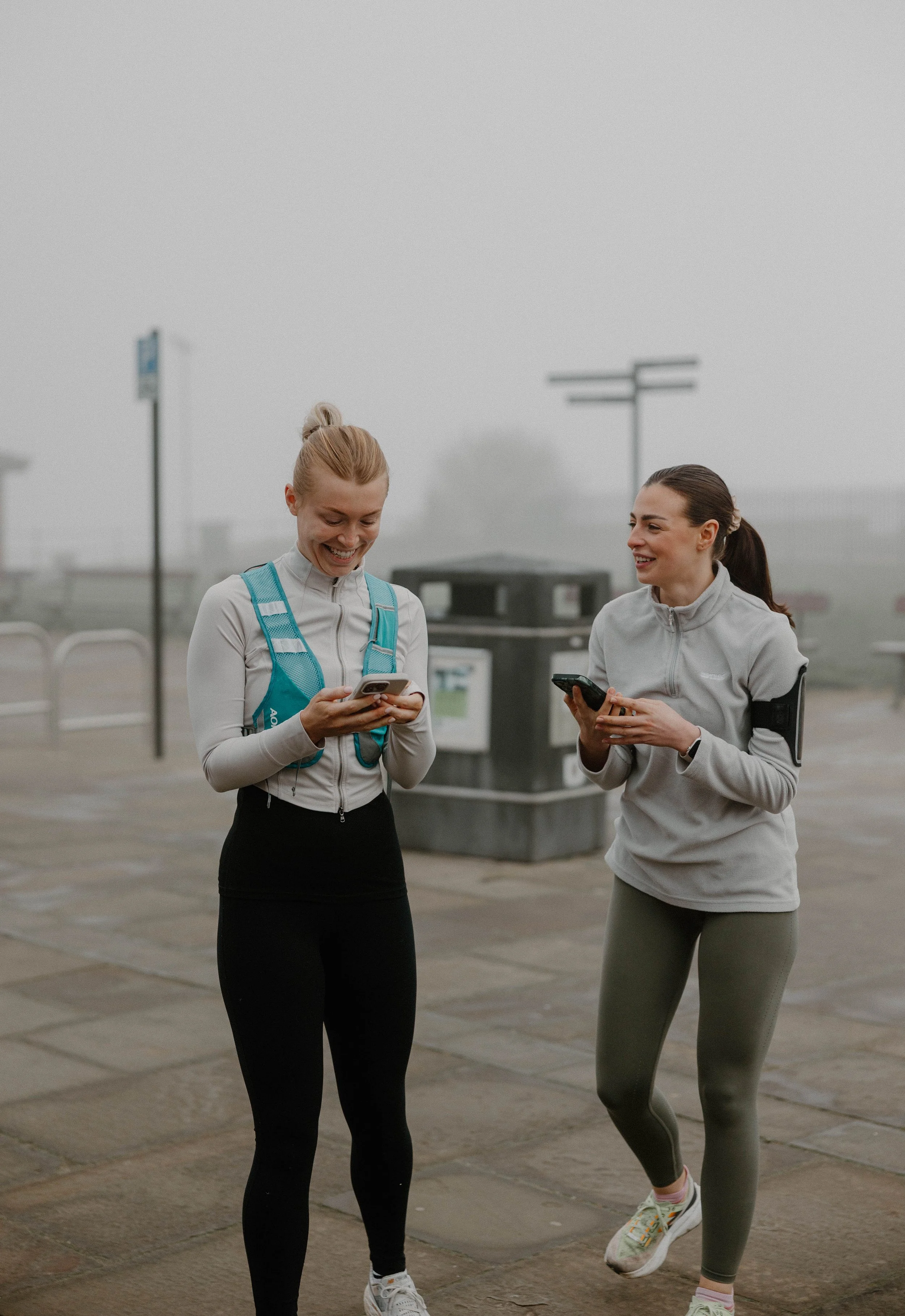 Two women in athletic wear smiling and looking at their phones outdoors on a foggy day.