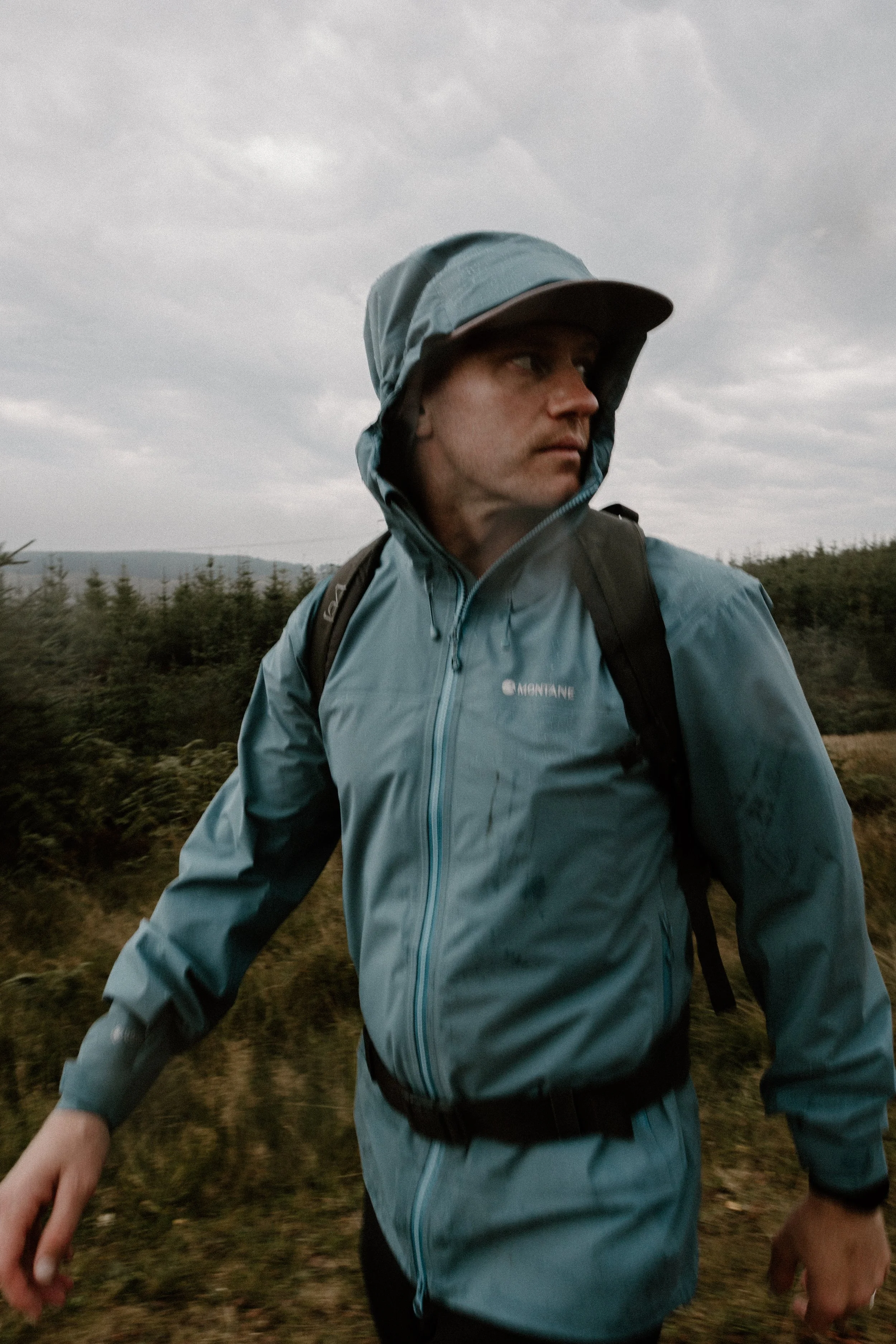 Man in a blue rain jacket with a hood, hiking outdoors with a backpack on a cloudy day.