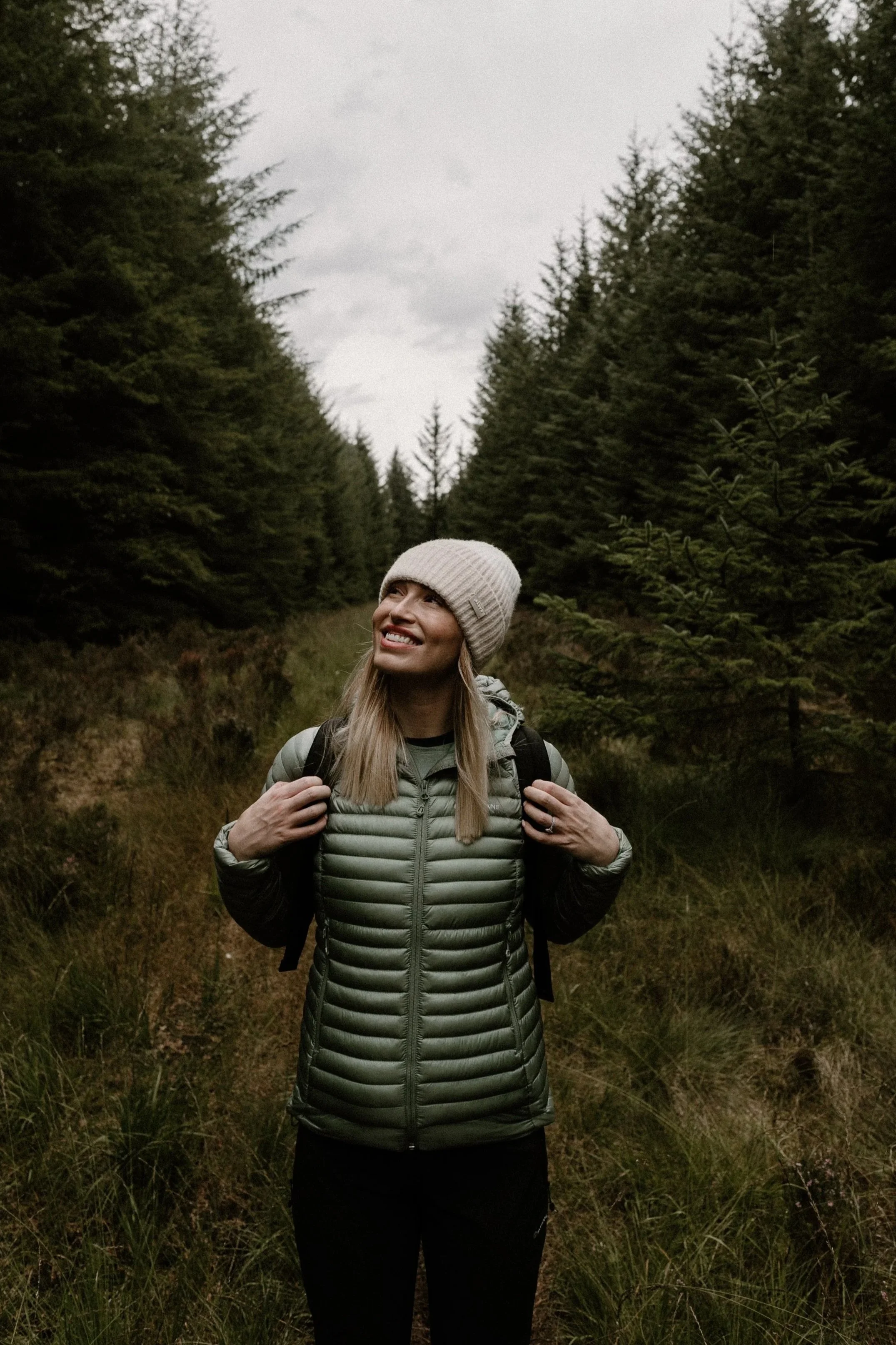 A woman smiling in a green puffer jacket and beige beanie, carrying a backpack, standing on a forest trail surrounded by tall pine trees.