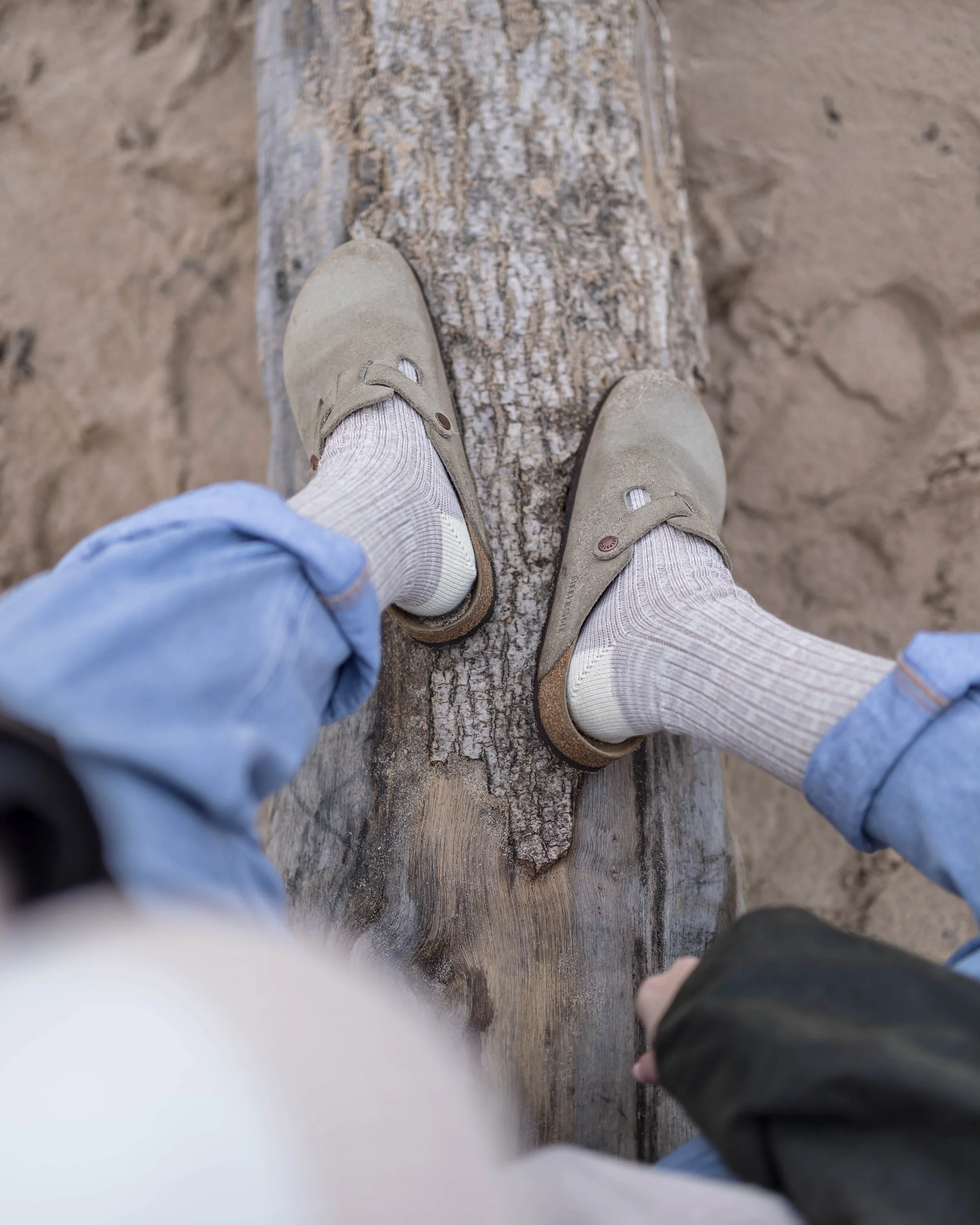 Person walking on a log at the beach, wearing beige shoes, cream-colored socks, blue jeans, and a black jacket.
