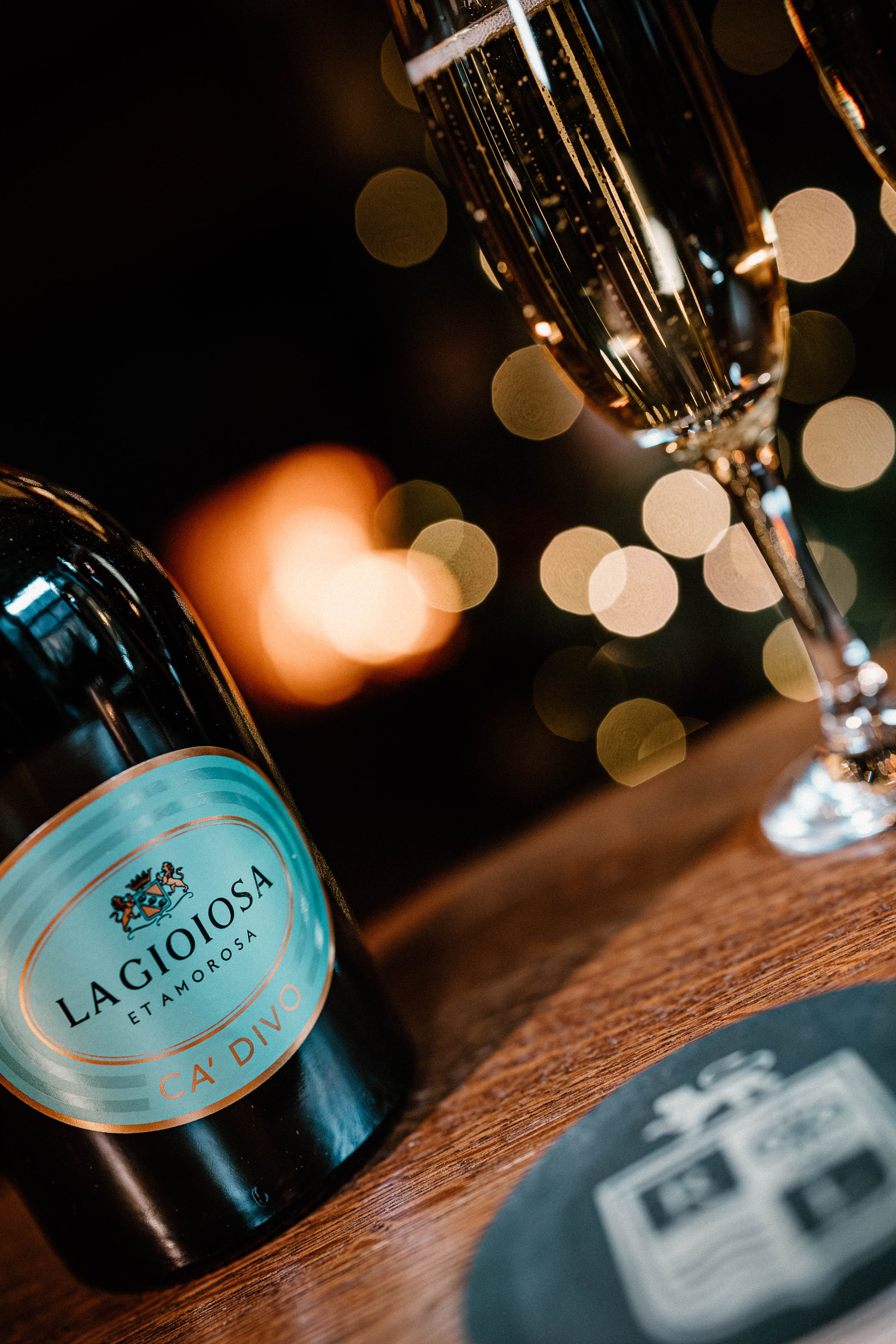 A bottle of La Gioiosa Etamorsa Ca' Divo sparkling wine and a filled wine glass on a wooden surface, with blurred lights in the background.