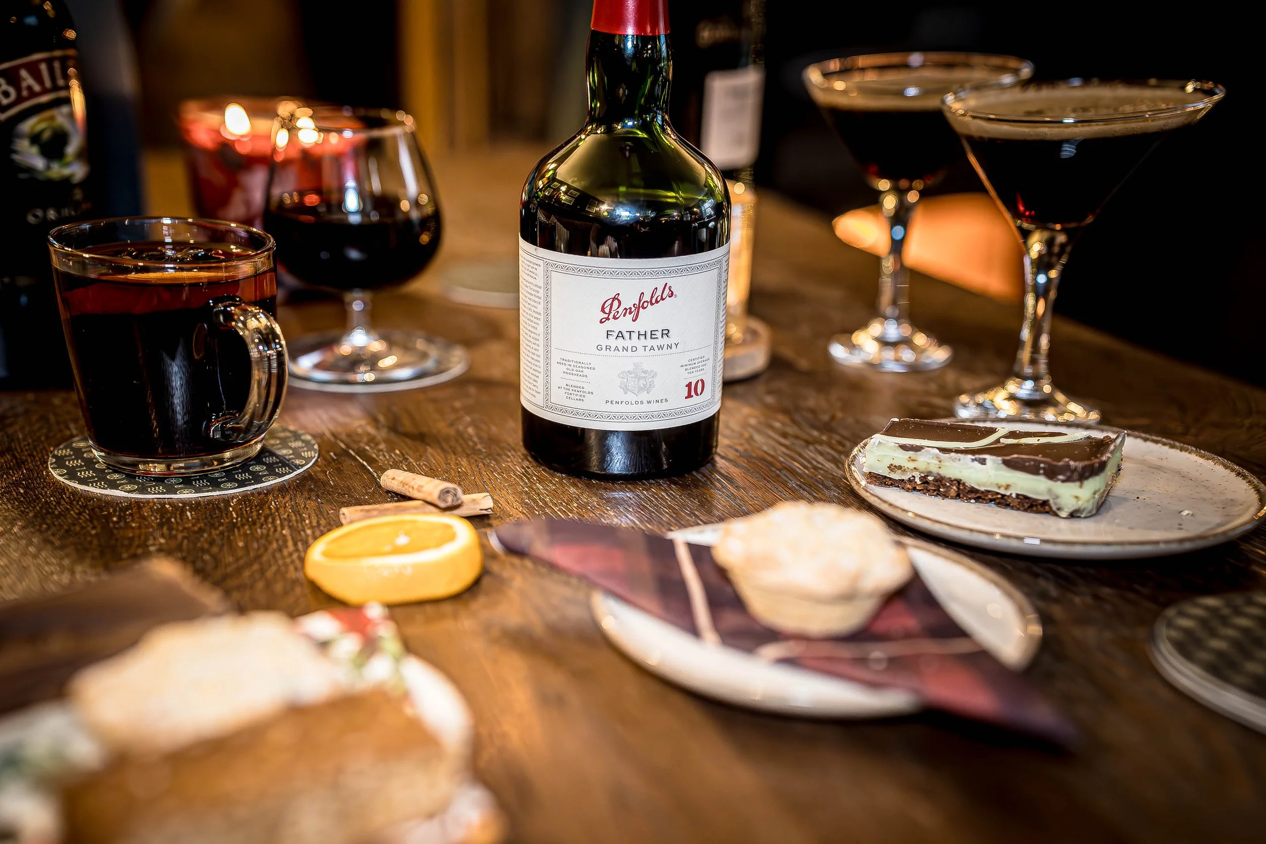 A wooden table set with glasses of red wine, a bottle of Penfolds Father wine, a slice of cheesecake on a plate, a lemon wedge, a cupcake, and some cinnamon sticks, with a blurred background.