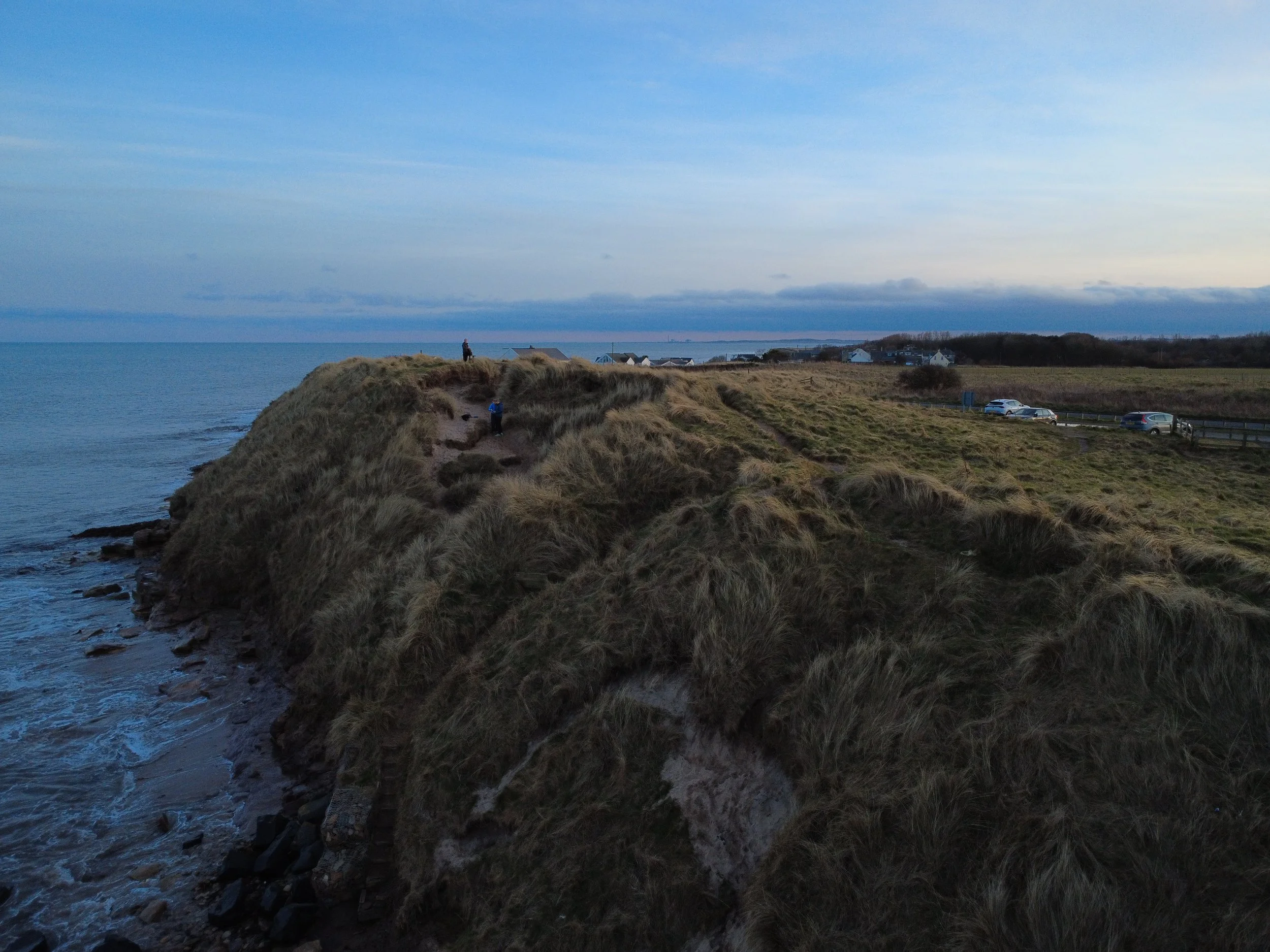 Cliffside with grassy terrain overlooking the ocean, with a few people walking along the path on the hilltop. There are parked cars and a small village in the distance under a cloudy sky.