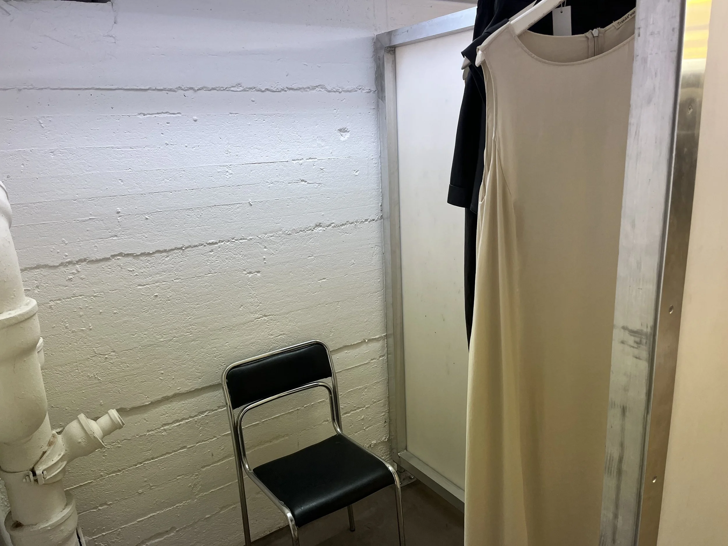 Ein schwarzer Stuhl steht neben einer weißen Ziegelmauer. Es gibt einen Wasserhahn auf der linken Seite und einen Umkleidebereich mit aushängenden Kleidern, darunter eine weiße, und einem schwarzen Kleid im hinteren Bereich.