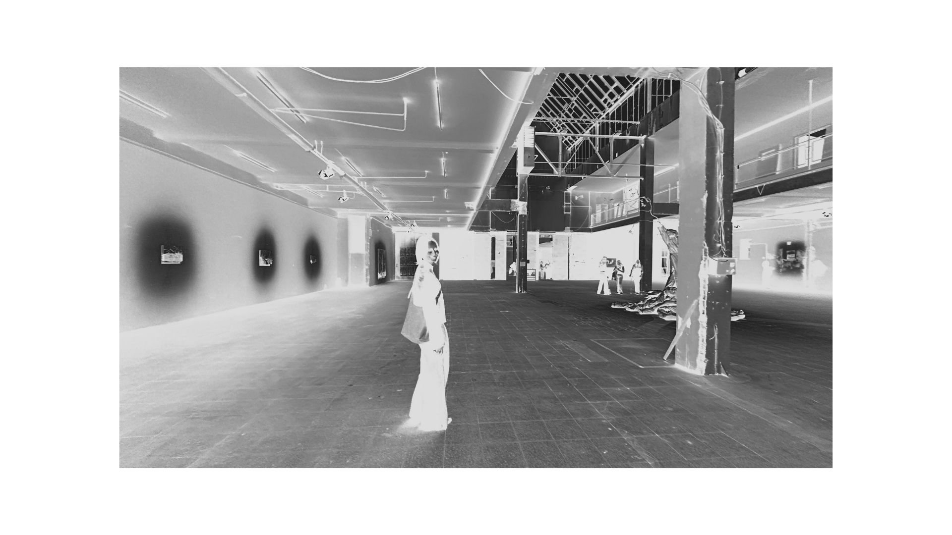 Ein leerer, moderner Innenraum mit einigen stehenden Menschen im Hintergrund, einem einzelnen stehenden Menschen im Vordergrund, und mehreren Bildern an der Wand auf der linken Seite. Das Bild ist in einer negativen Farbversion.