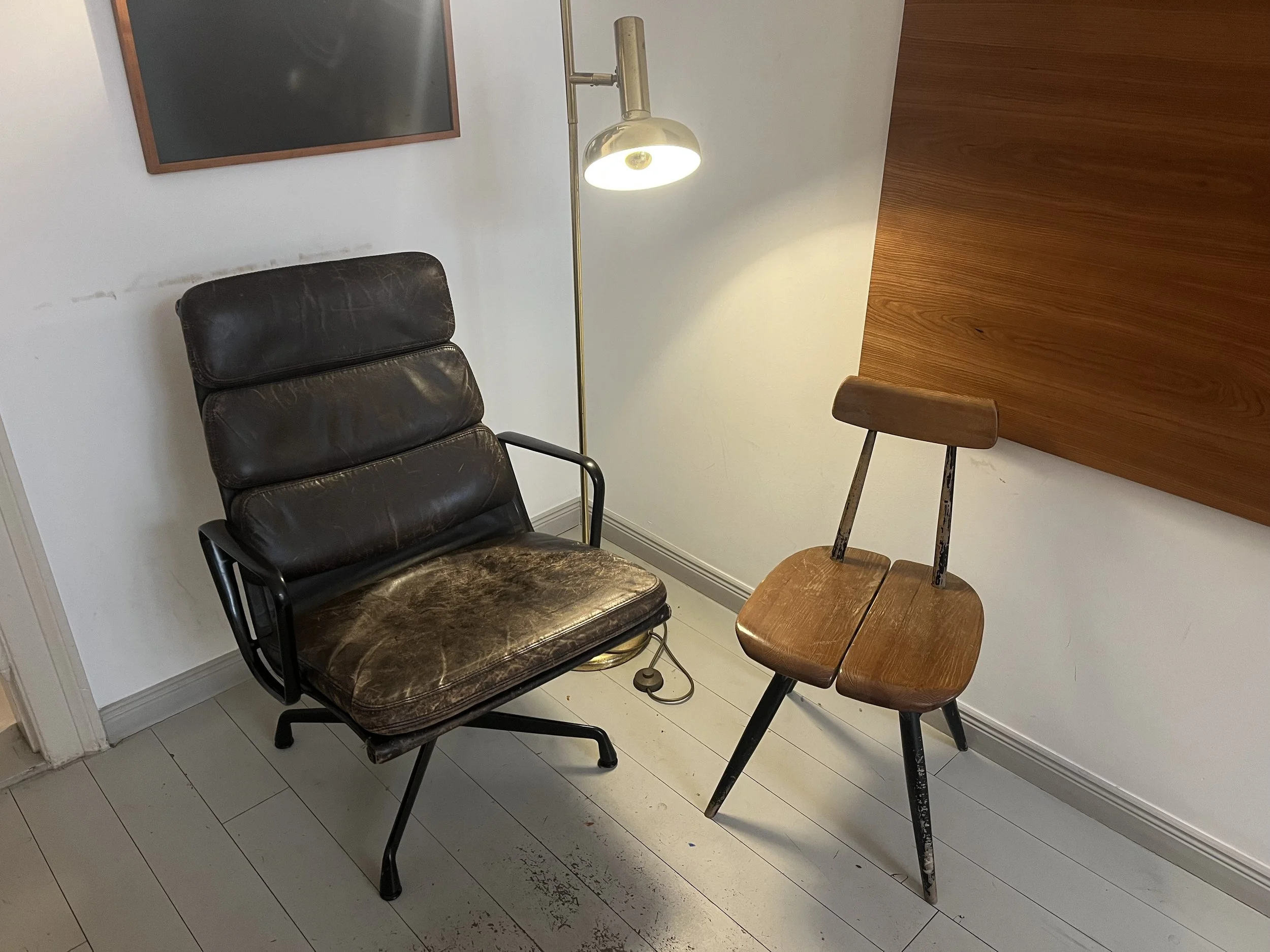 Ein alter, abgenutzter brauner Lederstuhl mit Armlehnen neben einem kleinen Holztisch. Daneben steht ein Stehlampe, im Hintergrund eine weiße Wand mit einem dunklen Bildrahmen.