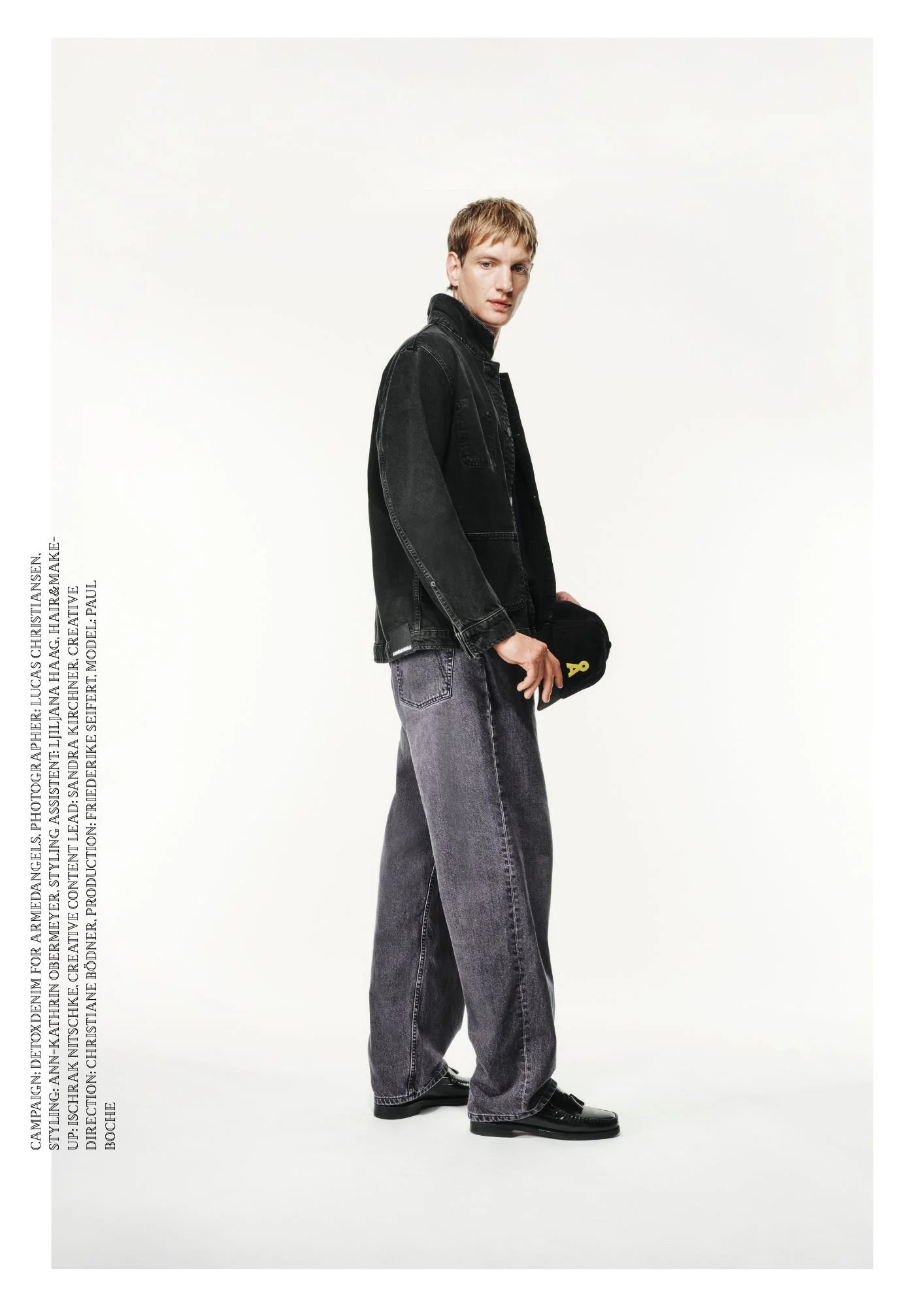 Modefotografie eines jungen Mannes in dunkler Jeansjacke, weiten grauen Jeans und schwarzen Schuhen, der eine schwarze Tasche hält, vor weißem Hintergrund.