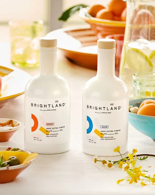 Two bottles of Brightland extra virgin olive oil, one labeled 'Awake' and the other 'Alive,' placed on a table with various bowls of food and a bowl of oranges in the background.