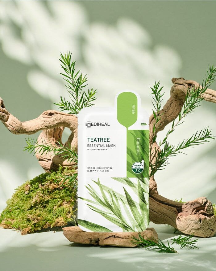 A package of MEDIHEAL Teatree Essential Mask, surrounded by green tea tree leaves and moss, with a wooden branch and a soft green background.
