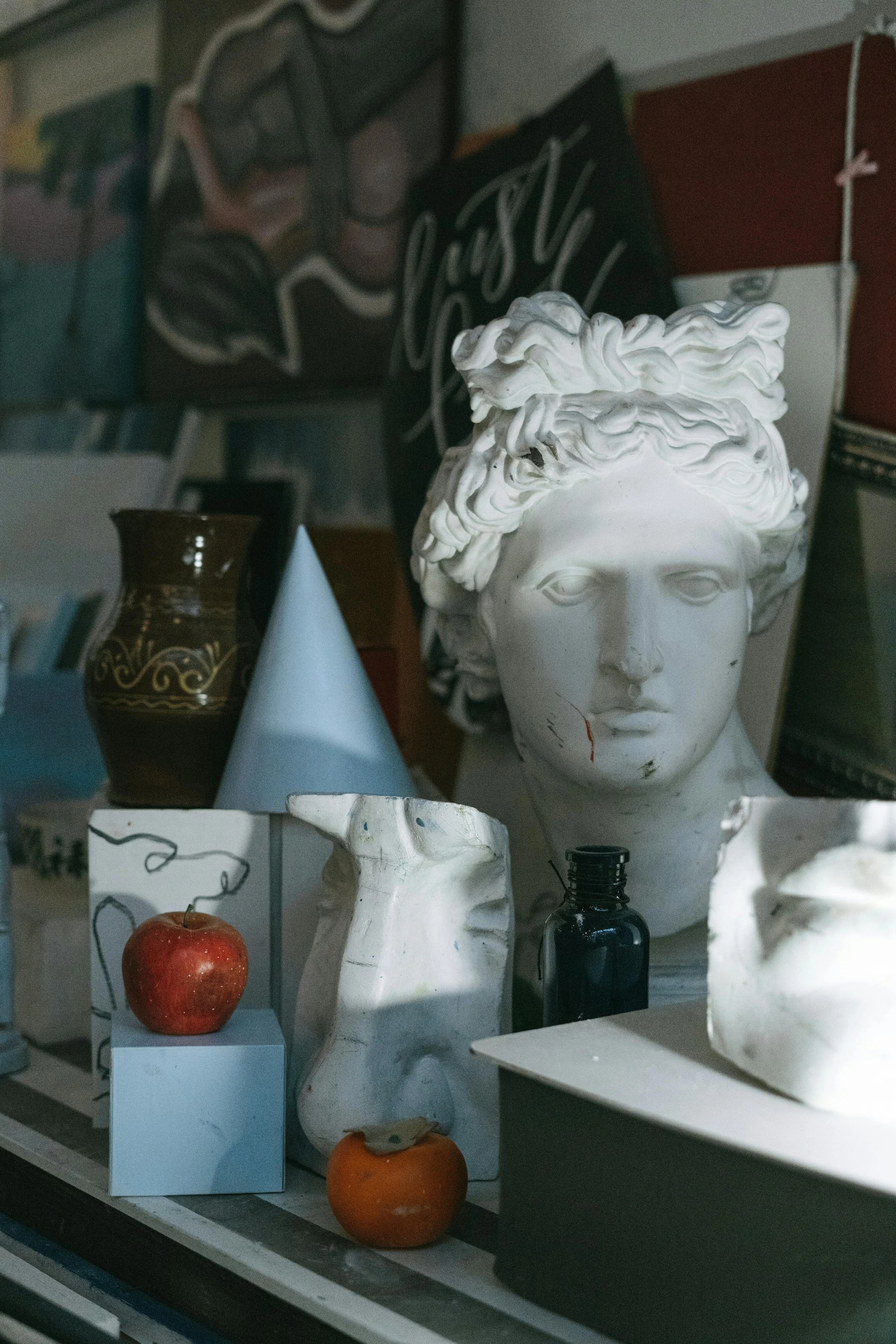 An art studio or display area featuring a classical bust sculpture, a dark vase with gold patterns, a light blue cone, an apple, a small orange, a black bottle, and various abstract and modern art pieces in the background.
