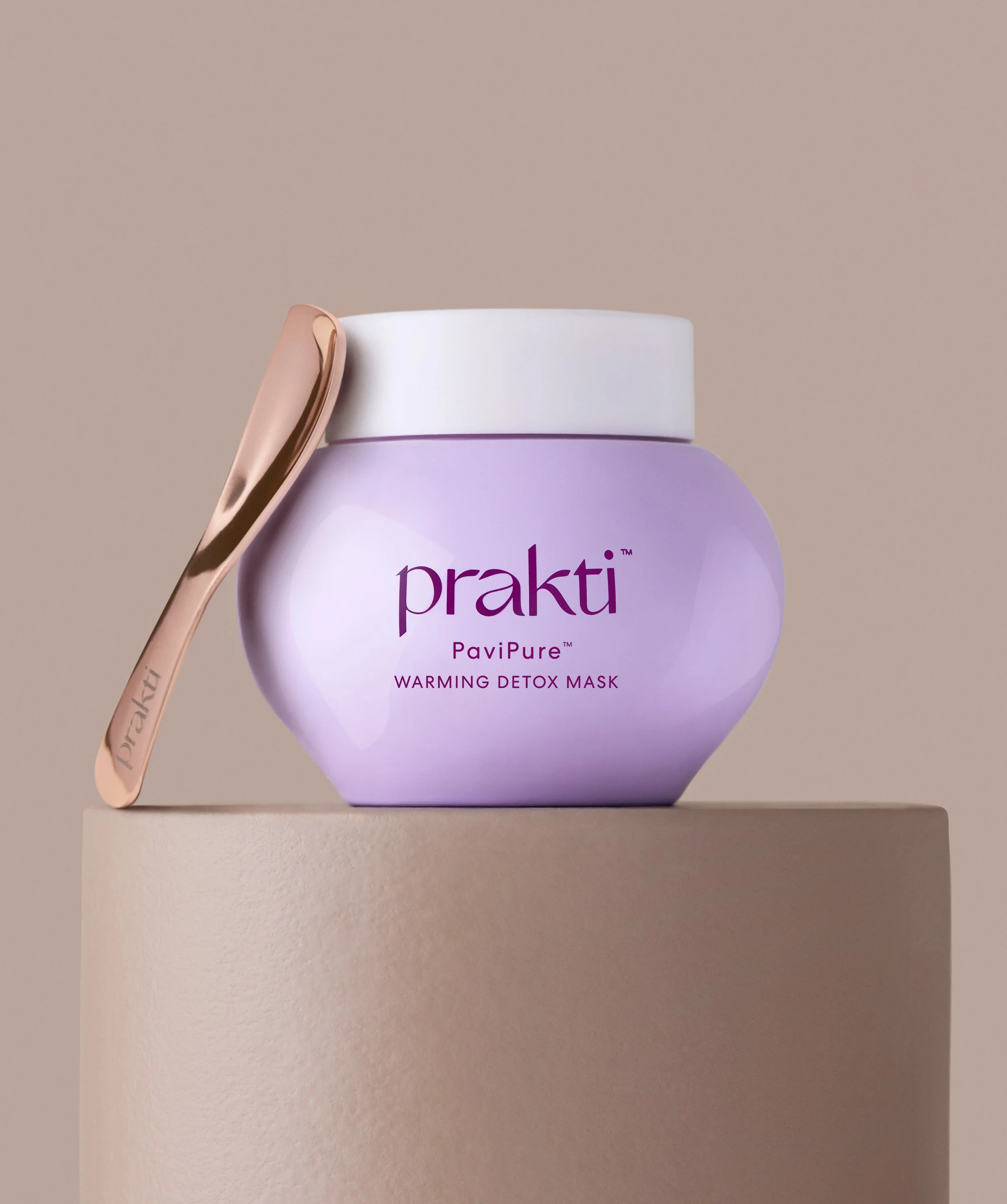 A purple jar of Prakti PaviPure Warming Detox Mask with a gold spatula leaning against it, placed on a beige pedestal against a plain background. A photo for marketing material for Metta Society.
