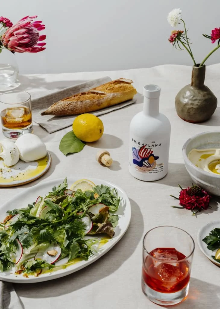 A table set with a variety of foods including a fresh green salad with lemon slices, a baguette bread, a plate of mozzarella cheese with olive oil, a glass of amber-colored drink with ice, a bottle labeled 'Brightland,' a bowl of hummus or dip with spoon, a vase with pink, white, and red flowers, and a glass of red drink. The table has a light tablecloth, and the background is white.