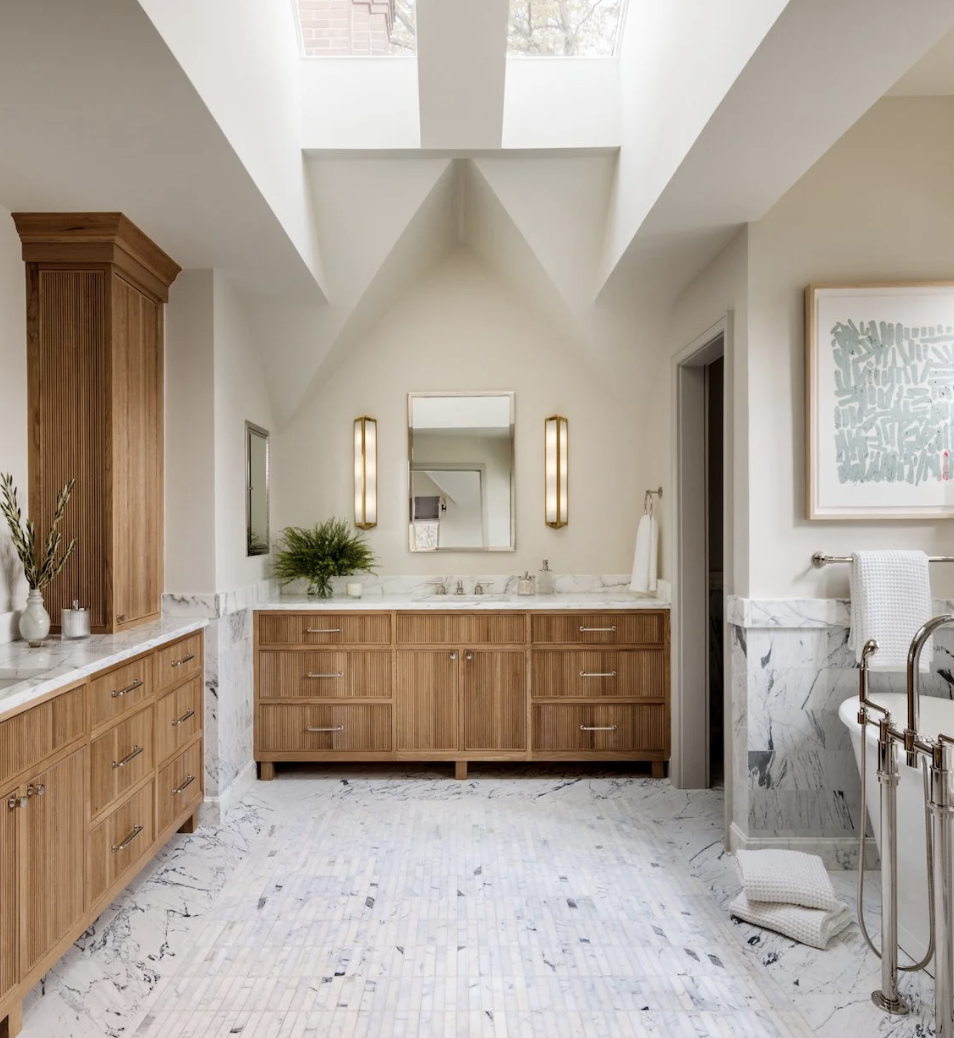 A bathroom with a skylight, wooden cabinets, marble floors, a mirror above the sink, and a bathtub with a towel hanging on the rail. Design by Emily Tucker Design.