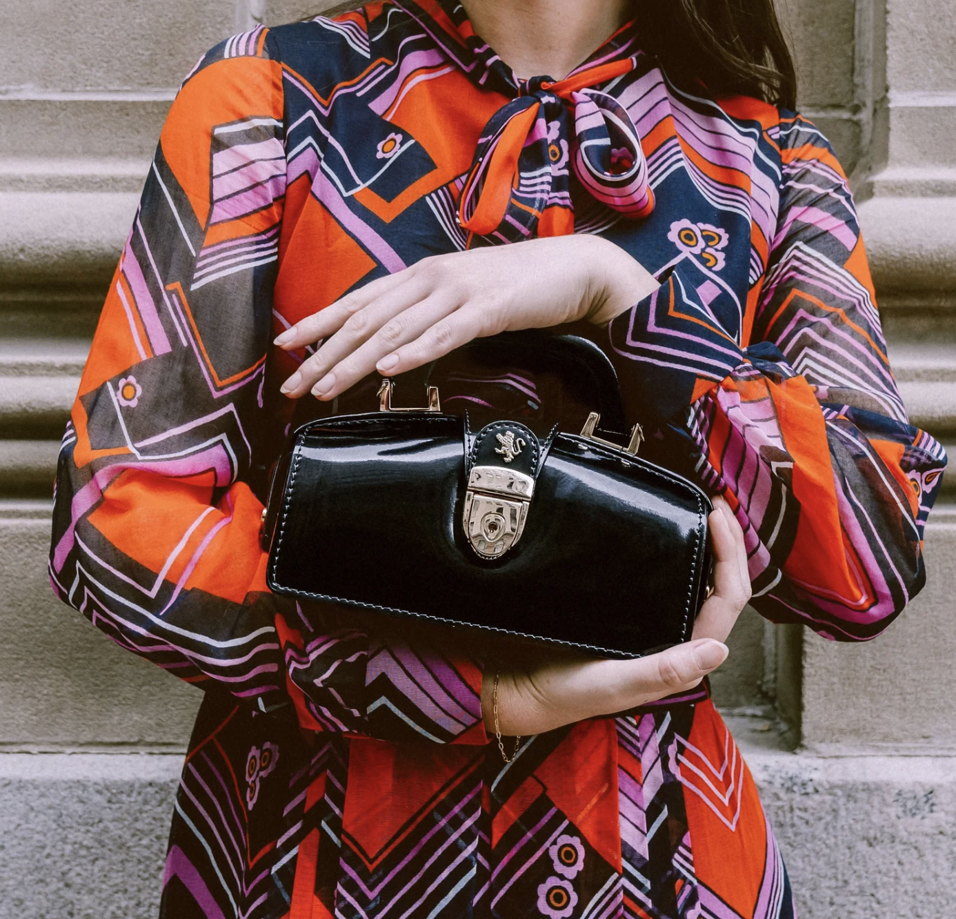 Person wearing a colorful geometric-patterned dress holding a shiny black handbag with a silver clasp, standing against a stone wall. Embassy London handbag for Metta Society marketing agency.