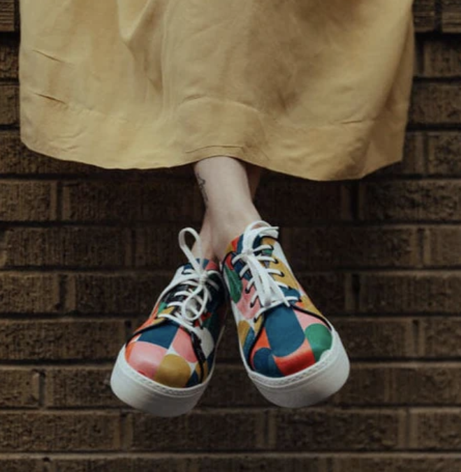 A person wearing a yellow skirt and colorful sneakers hanging above a brick wall. Embassy London shoes for Metta Society marketing agency.
