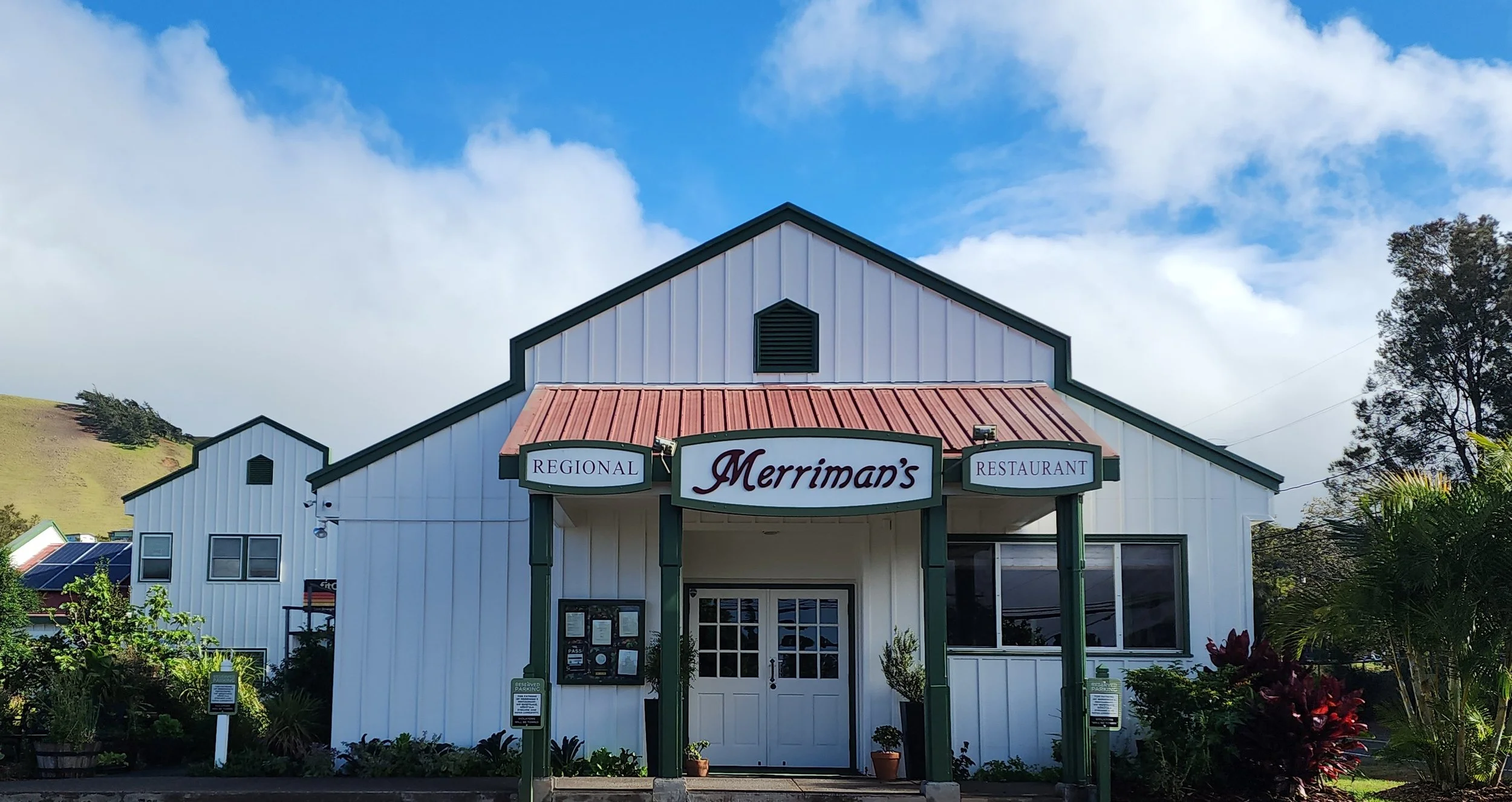 A white building with green trim and a red metal roof, with signs reading 'Regional Merriman's Restaurant' above the entrance, surrounded by plants and trees, under a partly cloudy sky.