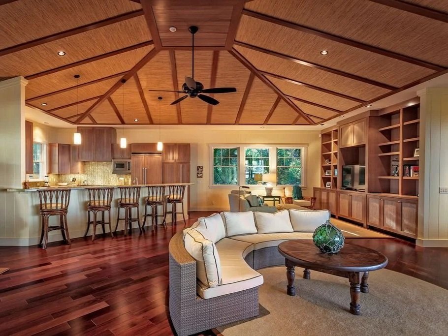 Open-concept living room with wood-paneled ceiling, wooden floor, cream-colored curved sofa, wooden coffee table with a green decorative object, and built-in wooden entertainment center with television. Kitchen area with barstools and pendant lightin