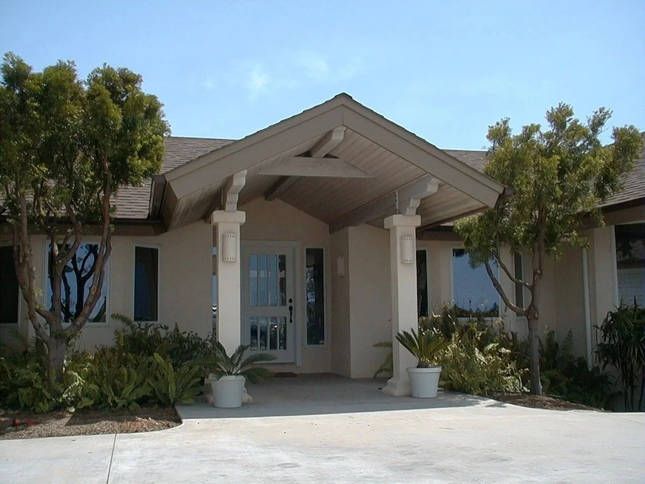 Front view of a modern house with a covered entrance, trees, potted plants, and a concrete driveway under a clear blue sky.