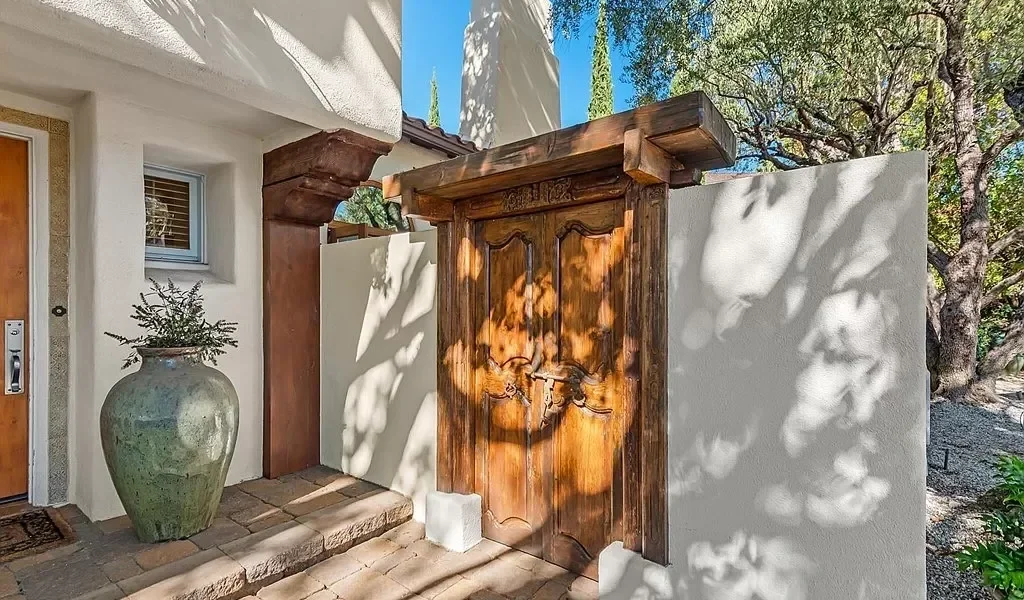 Wooden front gate with decorative carvings and metal latches, white stucco walls, large ceramic planter with greenery, stone pathway, and trees casting shadows on the wall.
