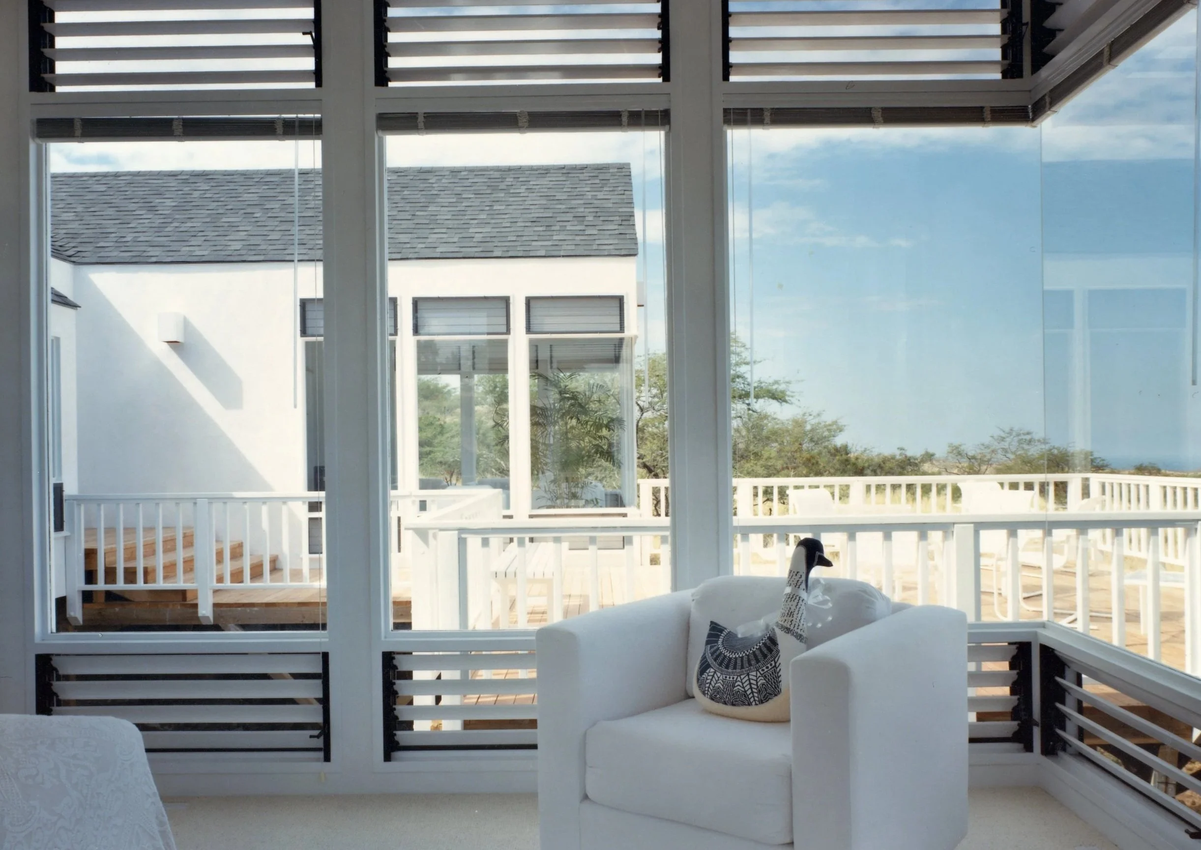Bright sunlit porch area with white railing and a glass enclosure. A white armchair with a decorative black and white duck pillow inside the enclosure. Outside, the porch has wooden decking, and the background shows a neighboring white house with mul
