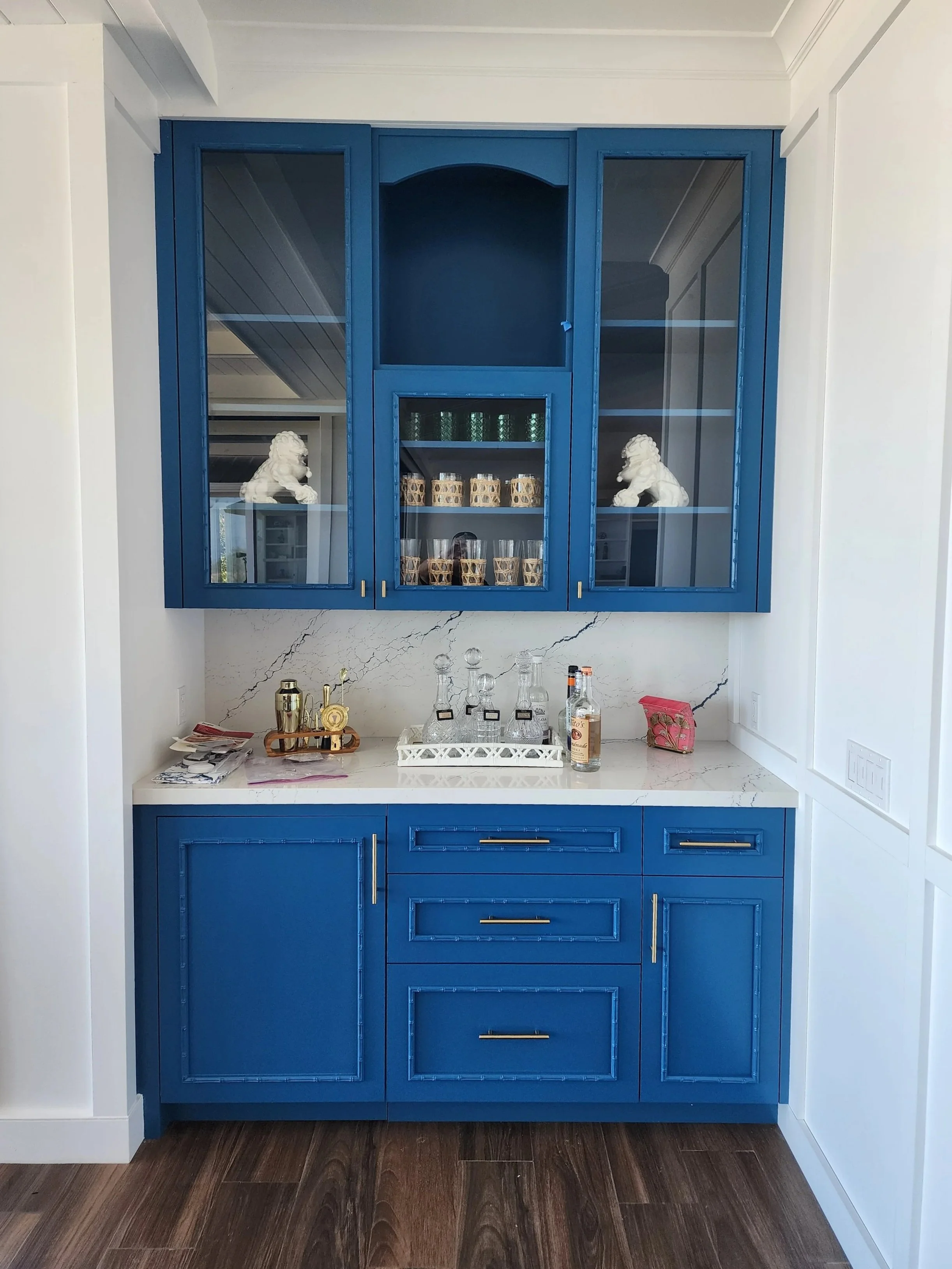 Blue bar cabinet with glass doors, white countertop, and barware. Decorative lion statues inside the cabinet, with glassware and bottles on the countertop. Dark wood flooring.