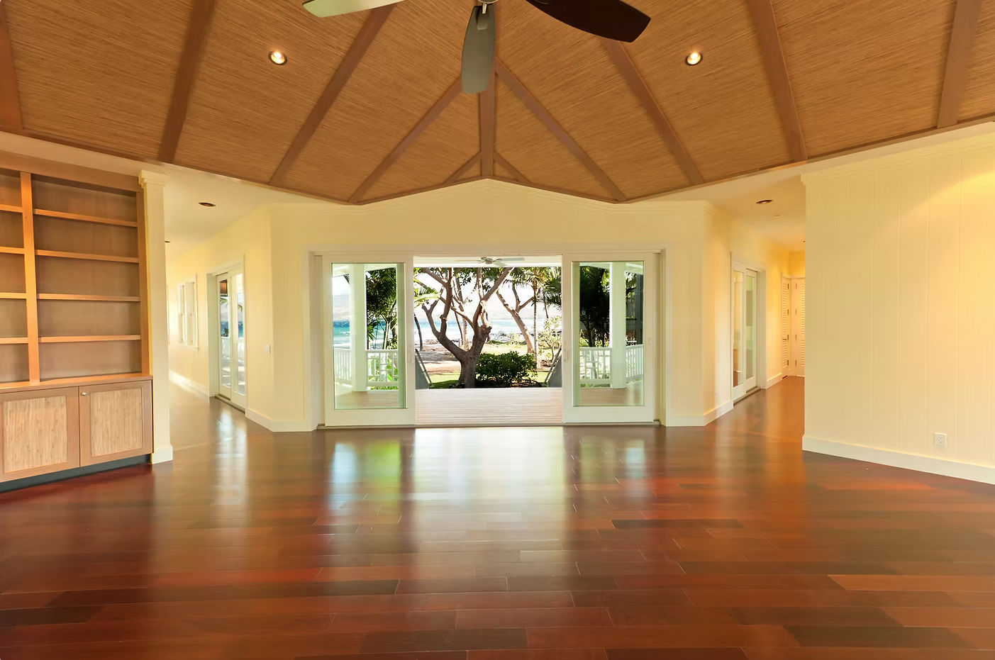 Empty living room with wooden floors, large glass sliding doors opening to a deck with trees and ocean view, ceiling fan, and built-in bookshelves.