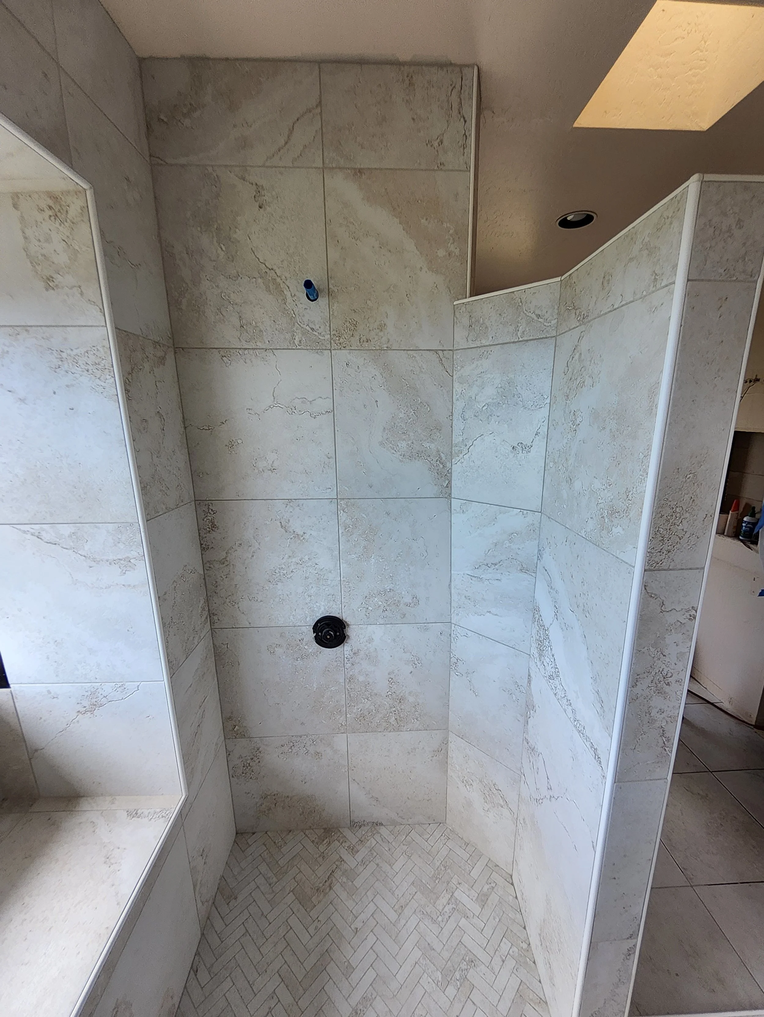 A bathroom shower area with beige marble tiles, an unfinished wall with pipe fittings for a showerhead and control, and a partial view of a built-in shelf.