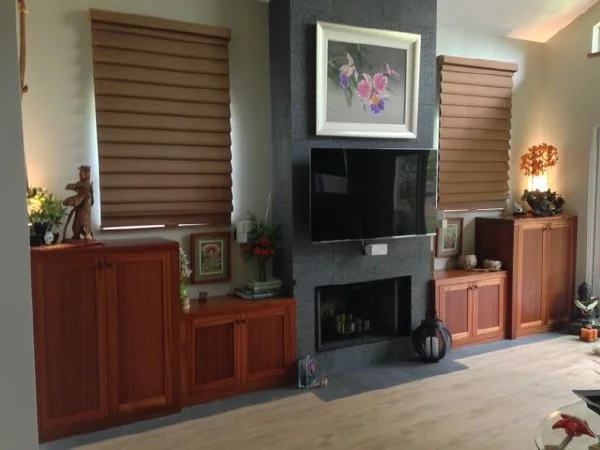 Living room with two wooden cabinets flanking a fireplace with a television above, framed floral artwork, window blinds, and decorative items.