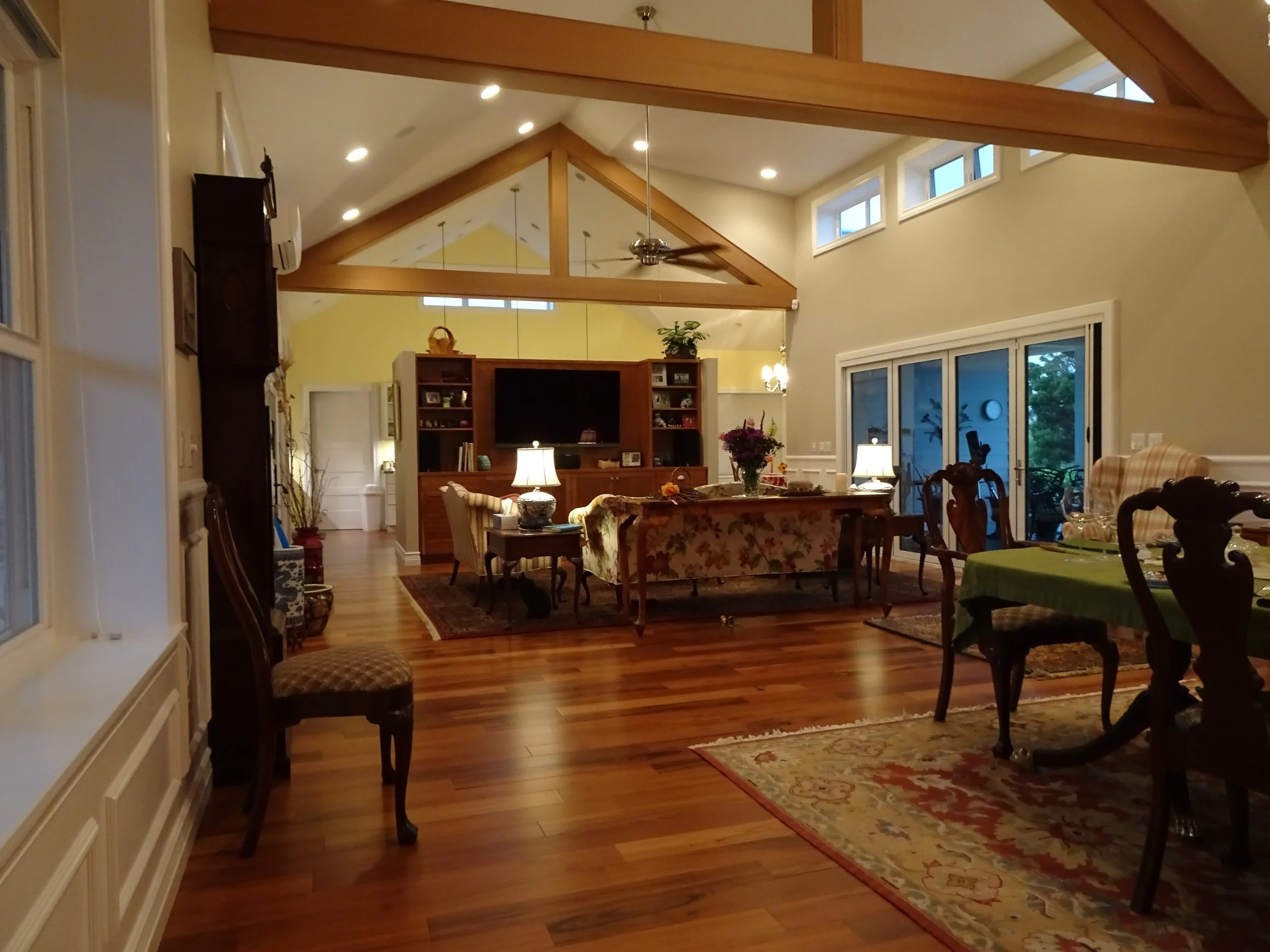 A spacious living room with high ceilings, wooden beams, hardwood floors, a large TV, multiple lamps, and floral furniture, illuminated by natural light from large sliding glass doors.