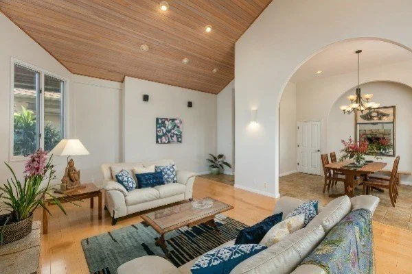 Living room with white sofas, blue cushions, wooden coffee table, potted plants, and a window with greenery outside, adjacent to a dining area with a wooden table and chandelier.