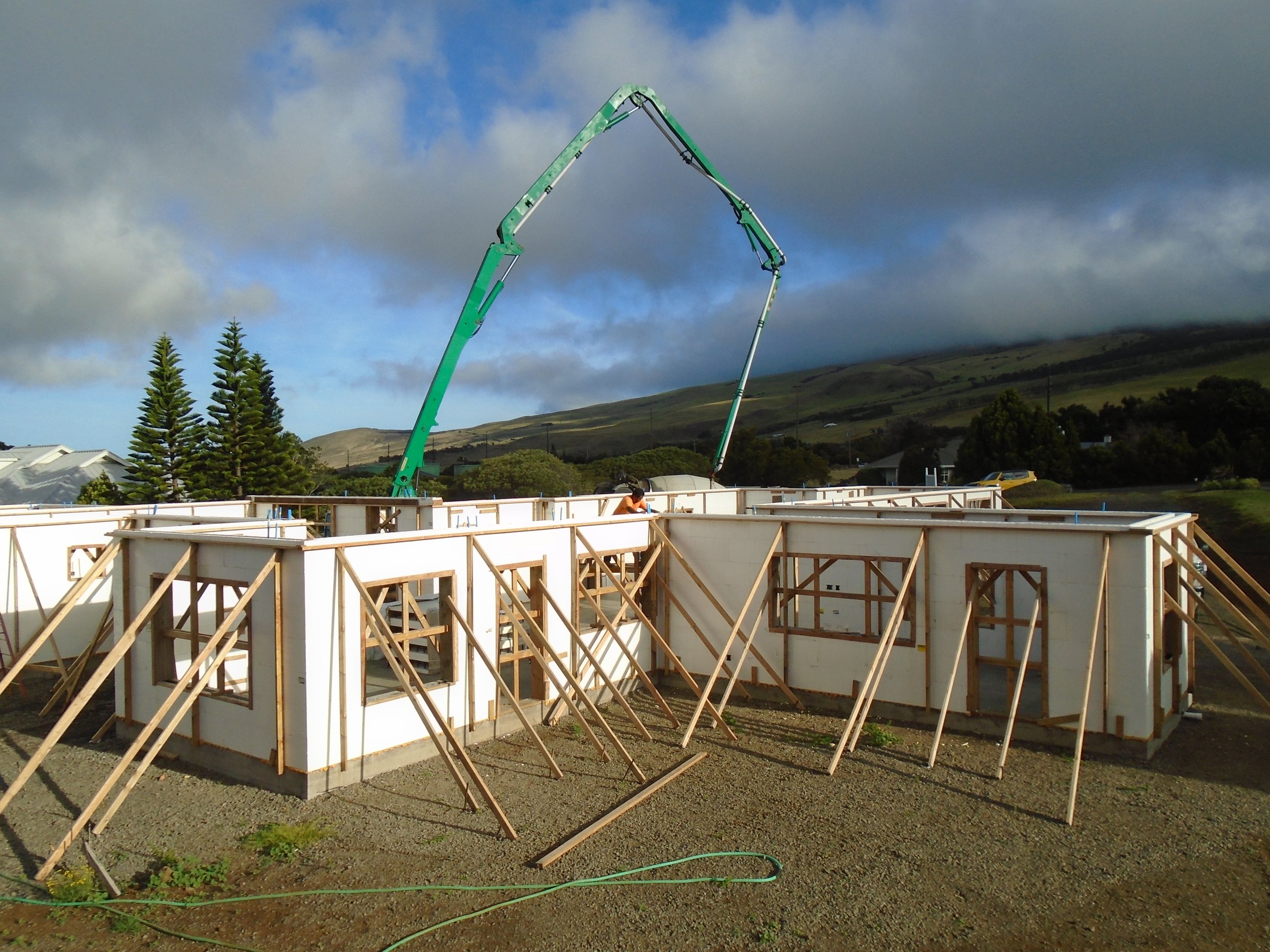 Construction site with wooden framing and a concrete pumping truck pouring concrete for building walls, surrounded by a rural landscape with trees and mountains in the background.