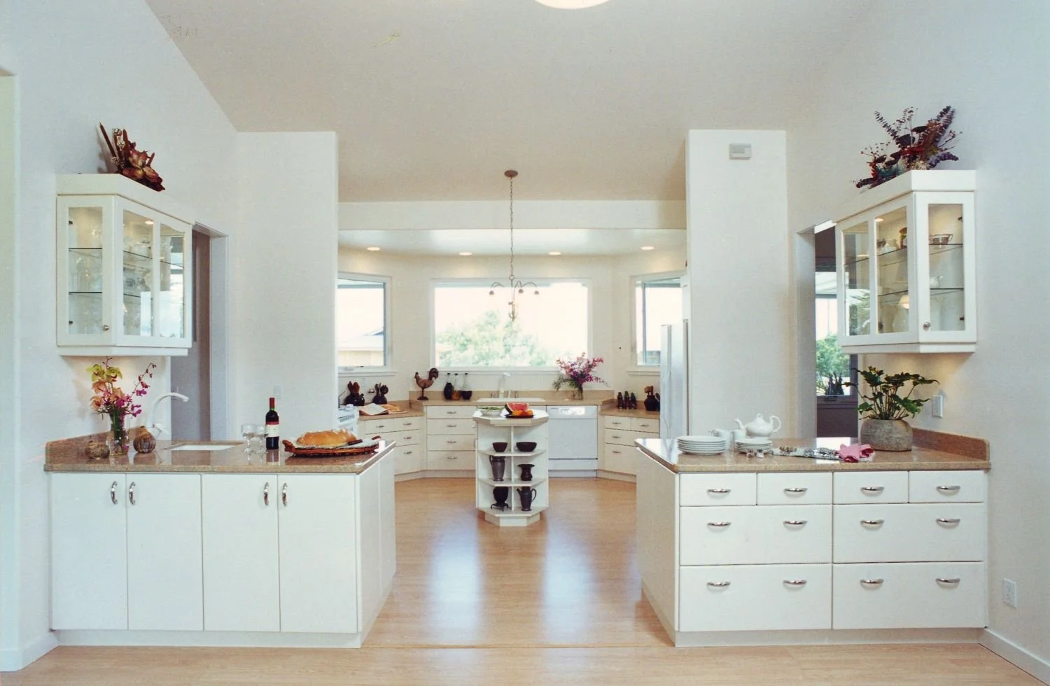 Bright kitchen with white cabinets, light wood floors, and multiple windows. Decor includes flowers and kitchenware.