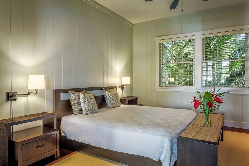 A bedroom with a bed, two nightstands, and two lamps on either side. The bed has decorative pillows and a white bedspread. There is a window with blinds and a view of greenery outside. A vase with red flowers and green leaves is on a wooden bench at 