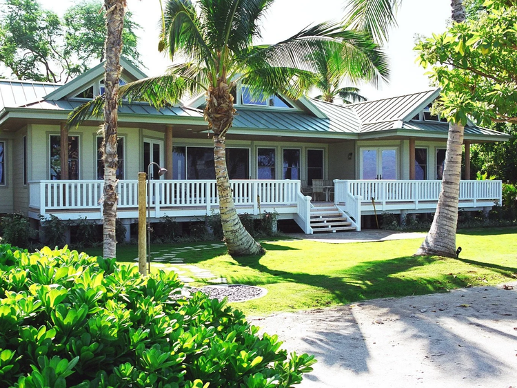 Big Island custom home exterior with green metal roof, wraparound white lanai, palm trees, and tropical landscaping.