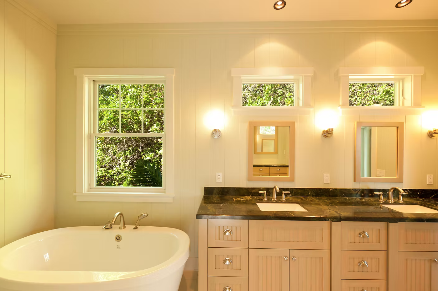 Bathroom with double vanity sinks, framed mirrors, wall sconces, a large window with greenery outside, and a corner bathtub.