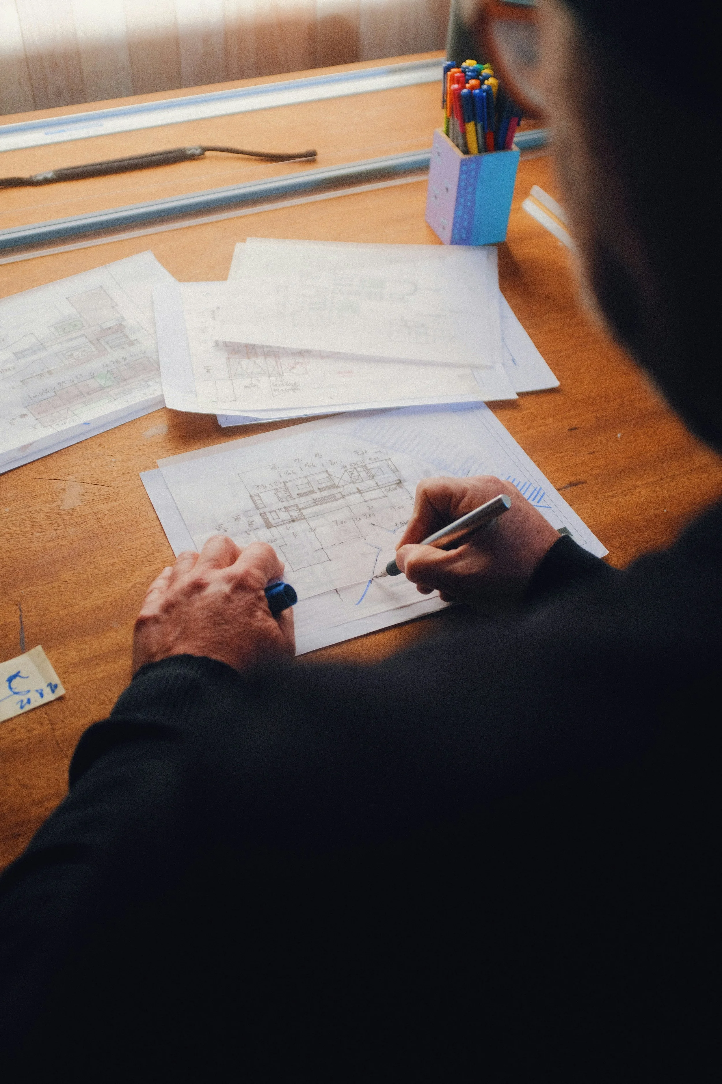 Person drawing architectural blueprints at a desk with papers, colored pens, and a ruler.