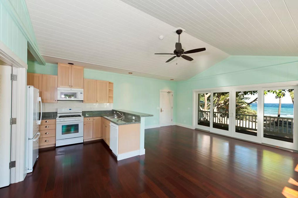 Living space with a kitchen, dark wooden floors, large windows showing a view of trees and the ocean, ceiling fan, and pastel green walls.