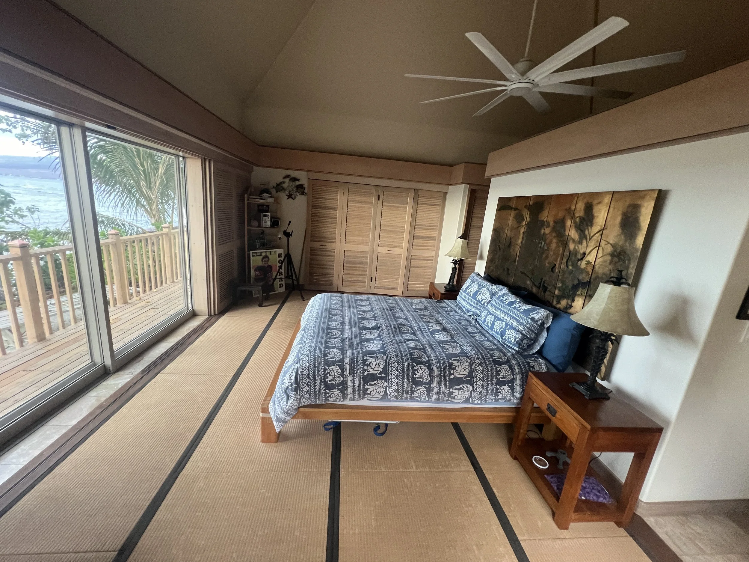 A bedroom with a wooden bed, blue patterned bedsheet, and matching pillow, beside a wooden nightstand with a lamp, in front of large glass sliding doors leading to a balcony overlooking water and palm trees, with a ceiling fan overhead.