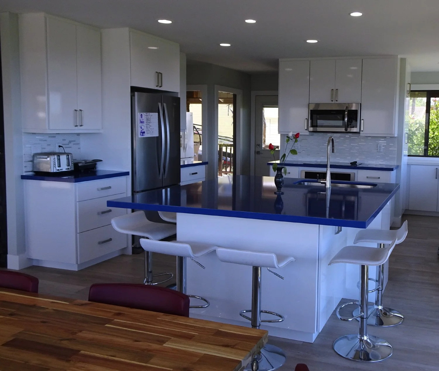 Modern kitchen with white cabinets, blue countertops, and a large island with a sink and four white bar stools. There is a vase with red roses on the island. Stainless steel appliances include a refrigerator, microwave, and toaster. Multiple ceiling 
